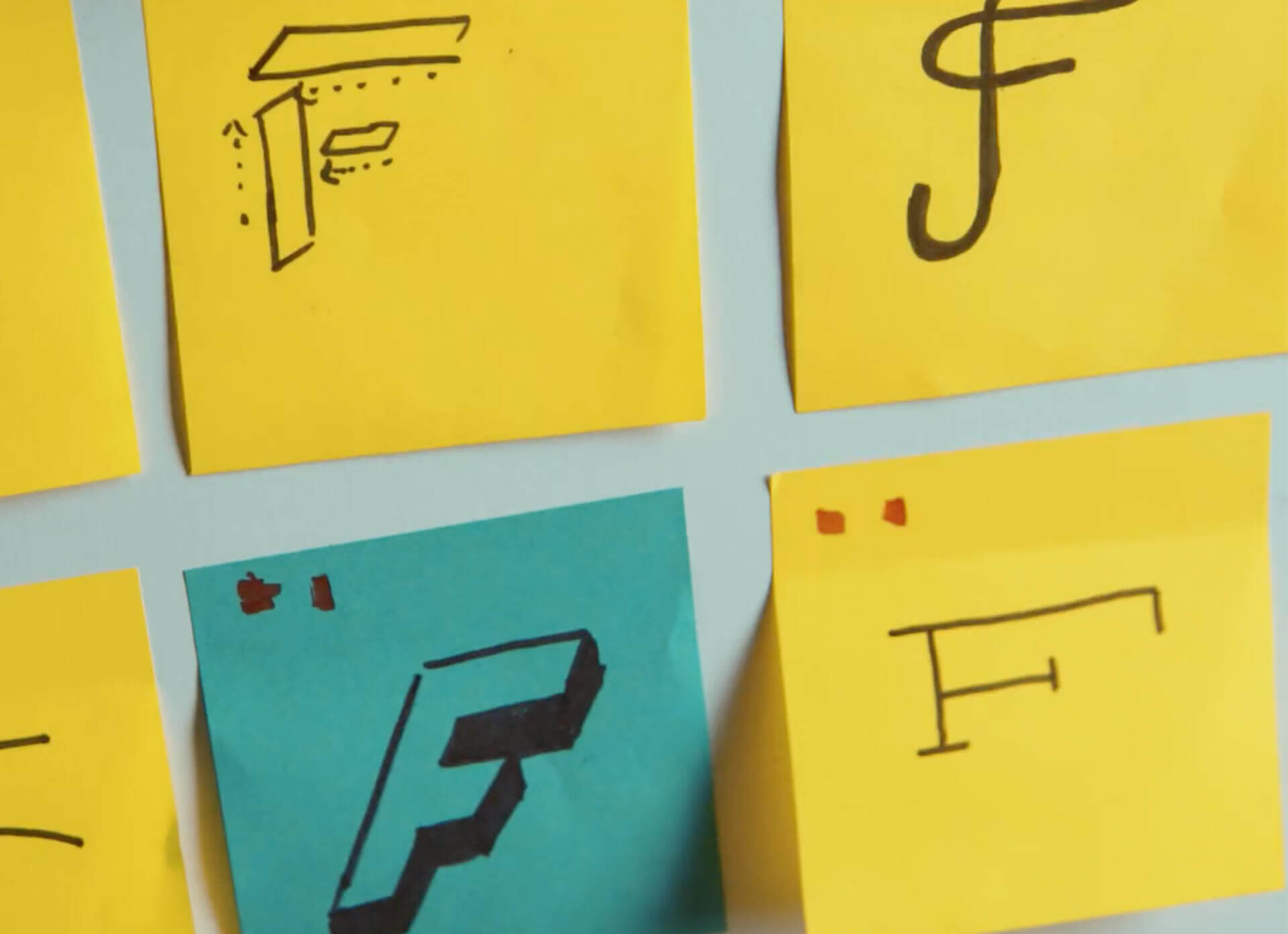 Four sticky notes, three yellow and one blue, display different hand-drawn letter F designs, each in a unique artistic style, arranged in a grid on a light surface.