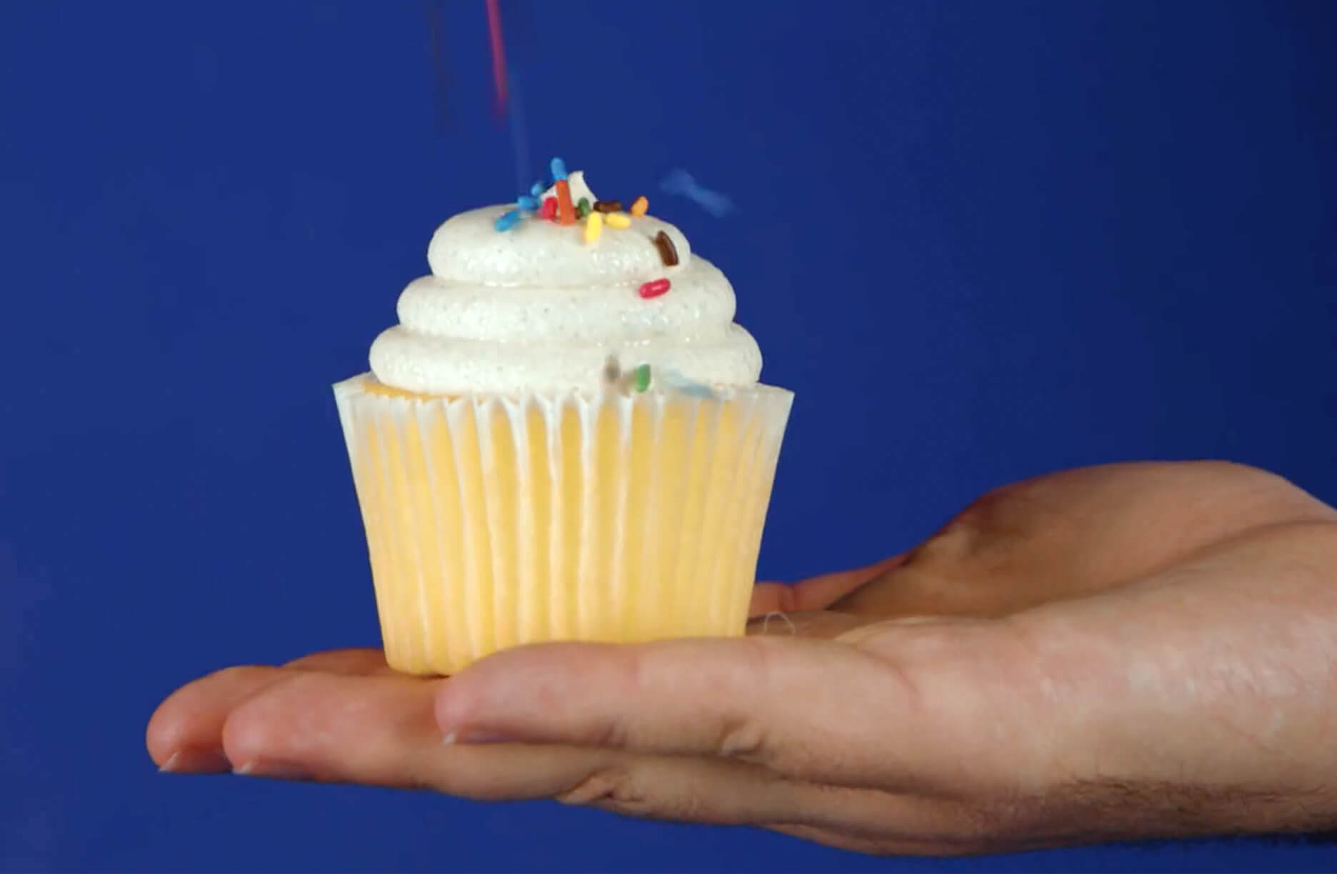 A hand holds a vanilla cupcake with white frosting and colorful sprinkles against a solid blue background.