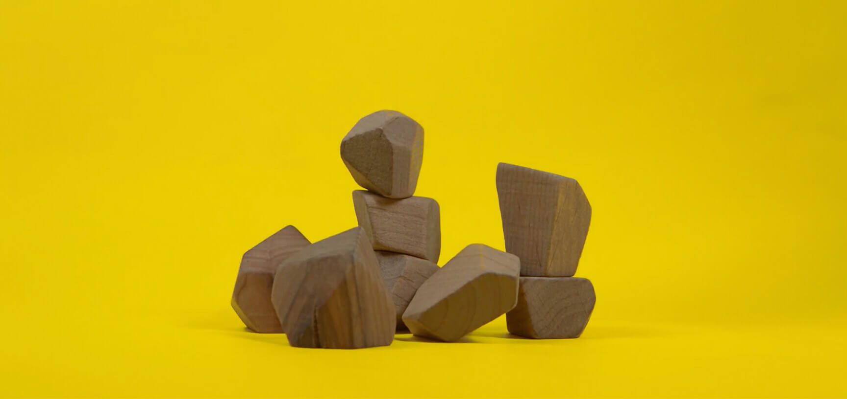 Seven irregularly shaped wooden blocks are stacked against a bright yellow background. Some blocks are balanced on top of others, forming a small, uneven structure.