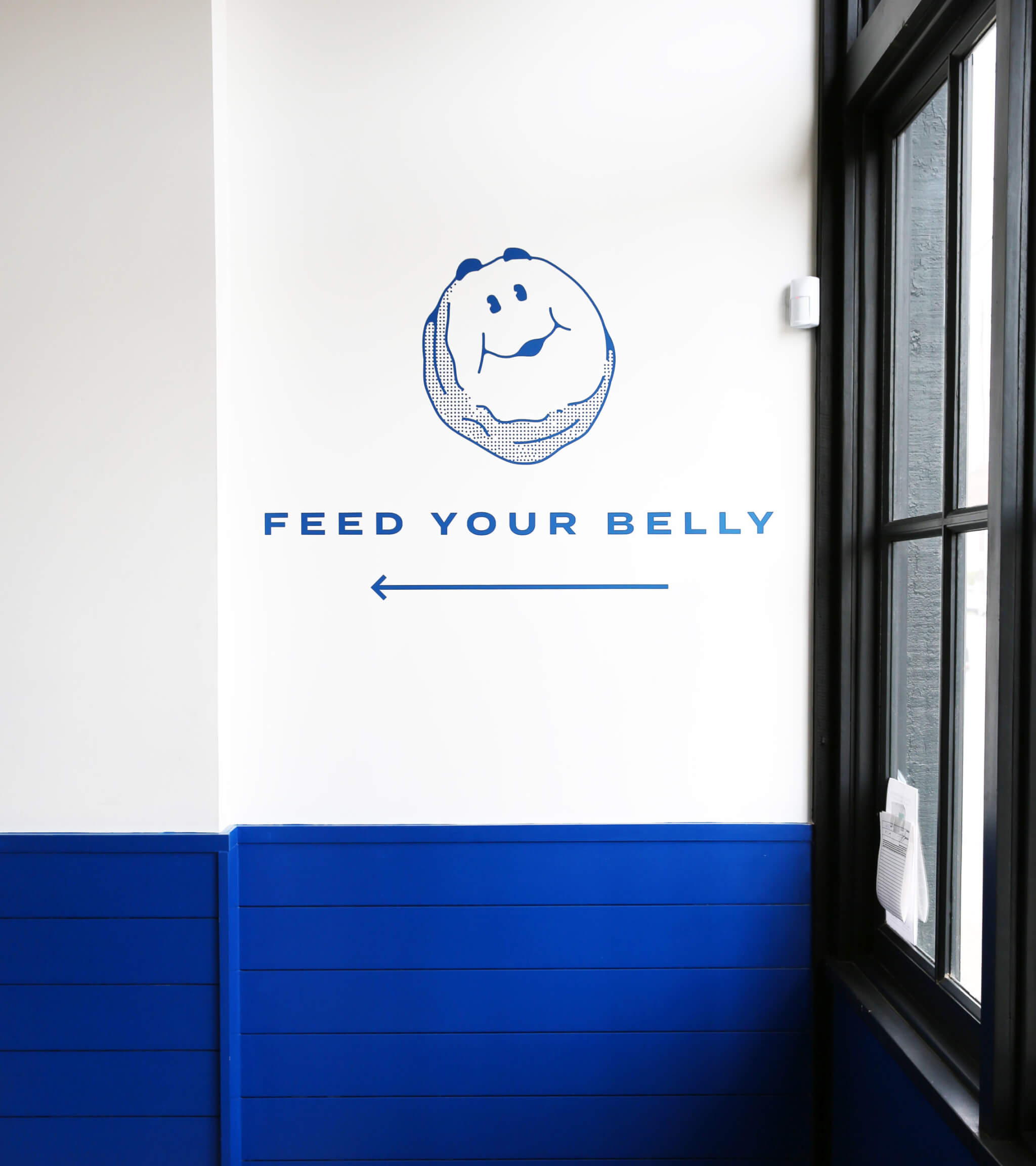 A white wall with a blue illustration of a smiling face above the words FEED YOUR BELLY and a left-pointing arrow. The lower part of the wall and window frame are painted blue.
