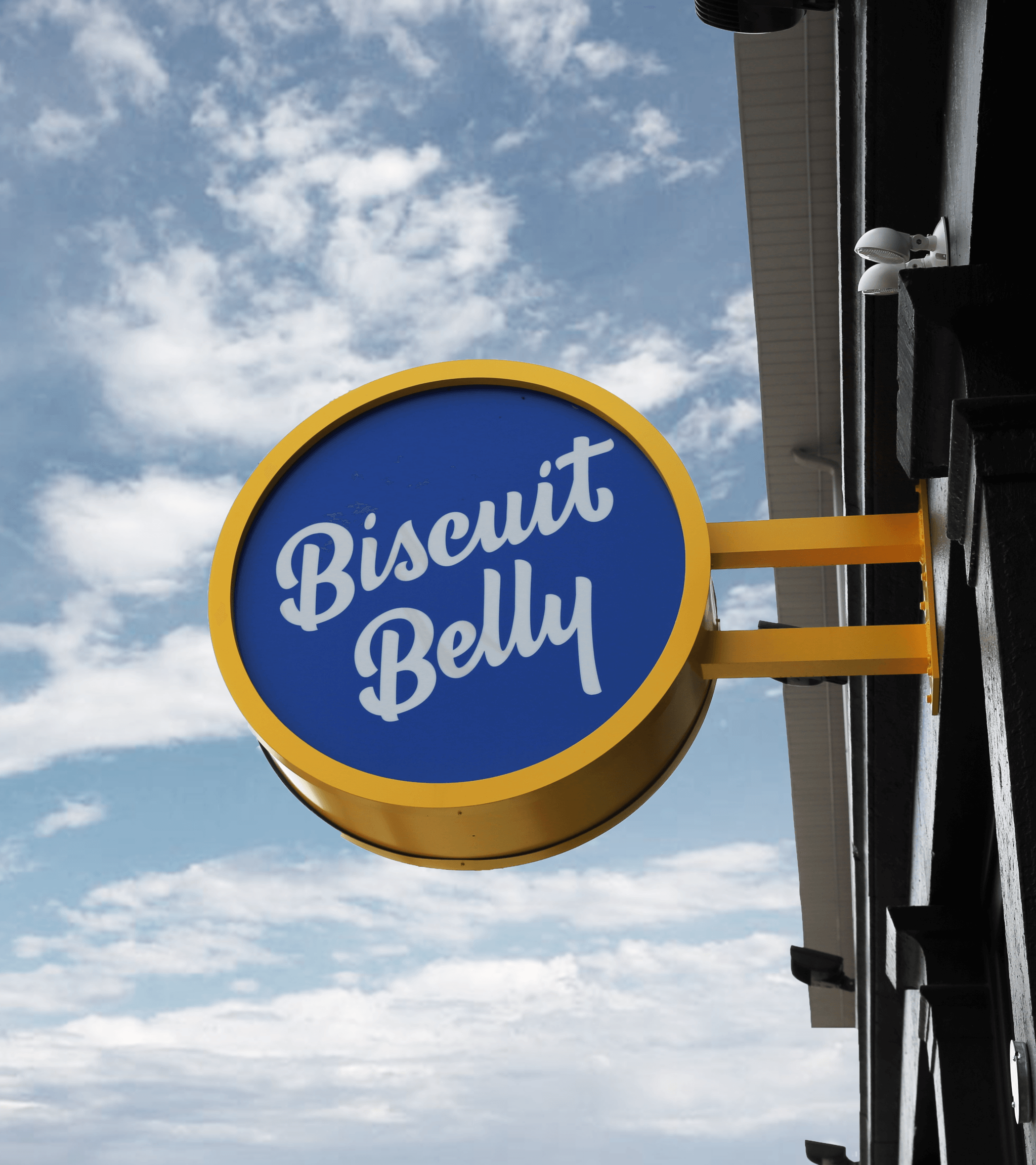 A round blue and yellow sign with white script text reading Biscuit Belly is mounted on the side of a building against a partly cloudy sky.
