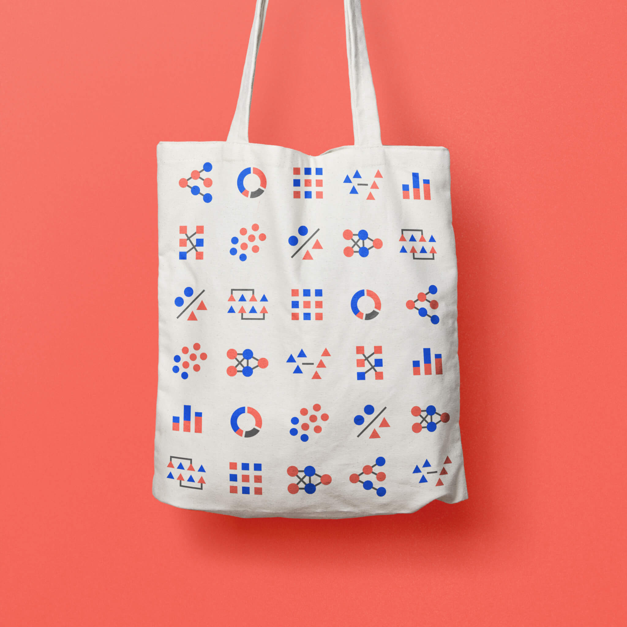 A white tote bag with colorful geometric patterns in blue, red, and gray, featuring circles, triangles, squares, and lines, hanging against a coral pink background.