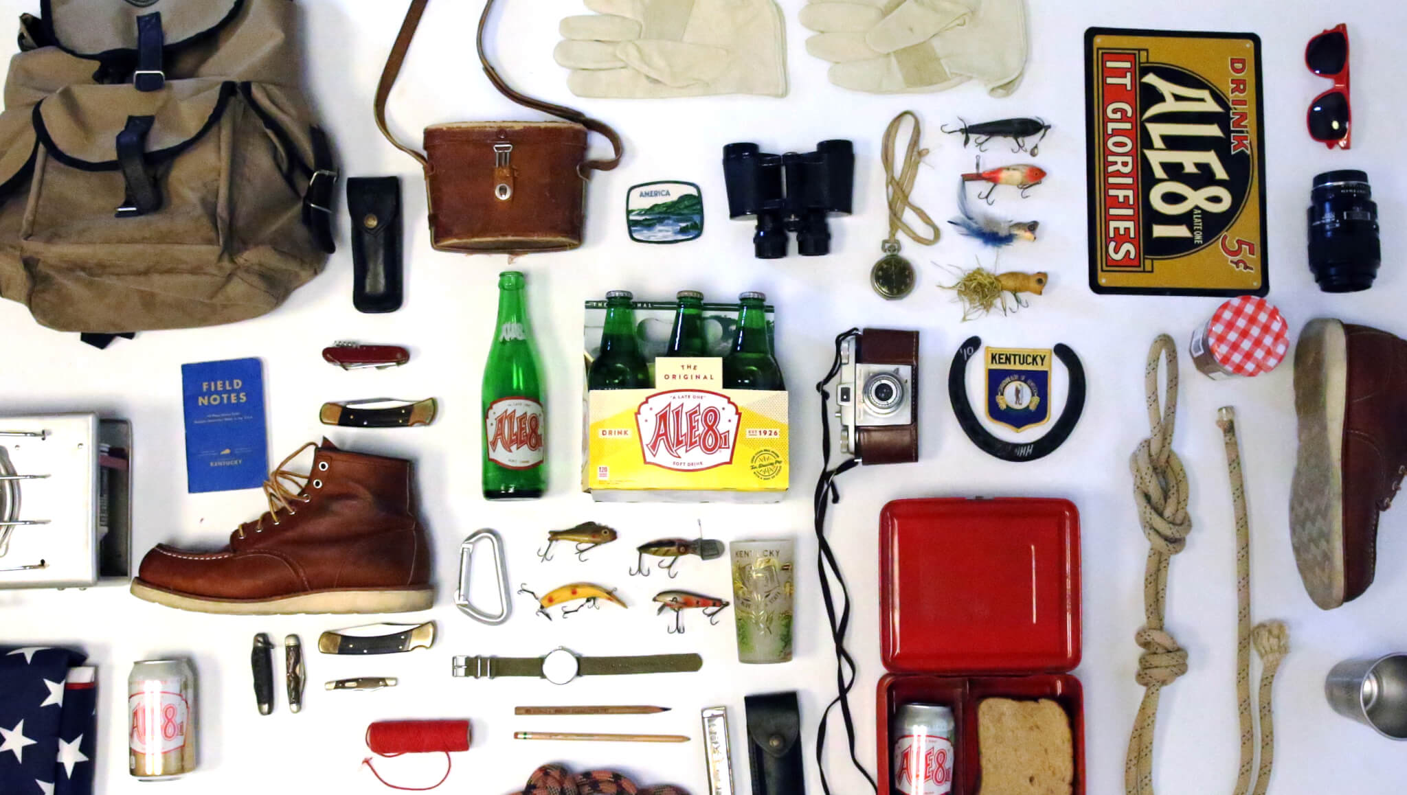A neatly arranged collection of vintage outdoor gear including boots, gloves, a backpack, camera, binoculars, fishing lures, rope, field notes, a watch, snacks, and Ale-8 soft drinks.