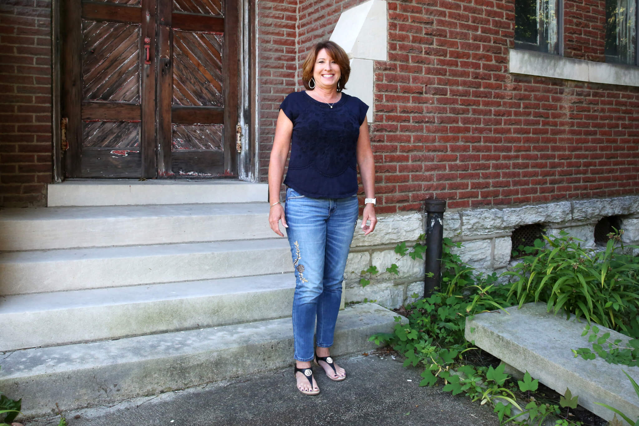 A woman wearing a navy shirt, jeans, and sandals stands smiling in front of a brick building with stone steps and greenery nearby.