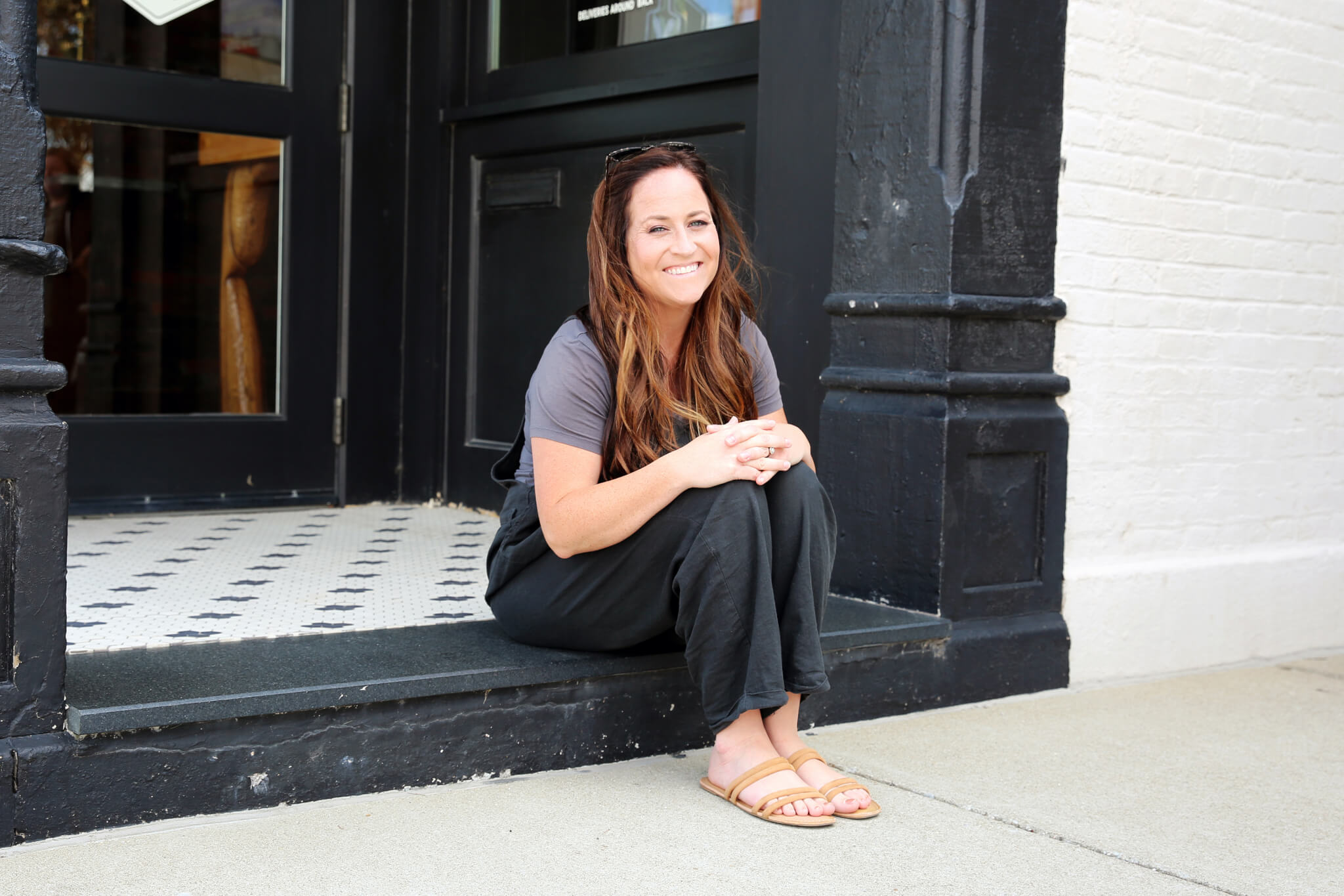 A woman with long brown hair, wearing a gray t-shirt, black pants, and sandals, sits smiling on a doorstep outside a building with black trim and a tiled entrance.