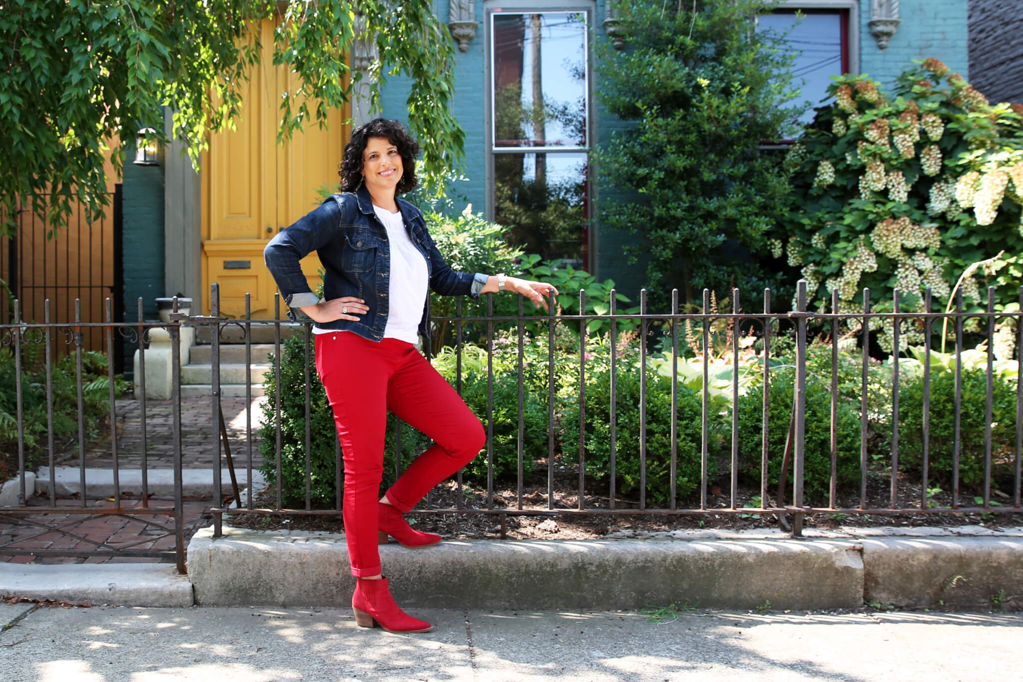 A woman with curly dark hair wearing a denim jacket, white shirt, red pants, and red boots stands smiling by a black metal fence in front of a house with a yellow door and lush green plants.