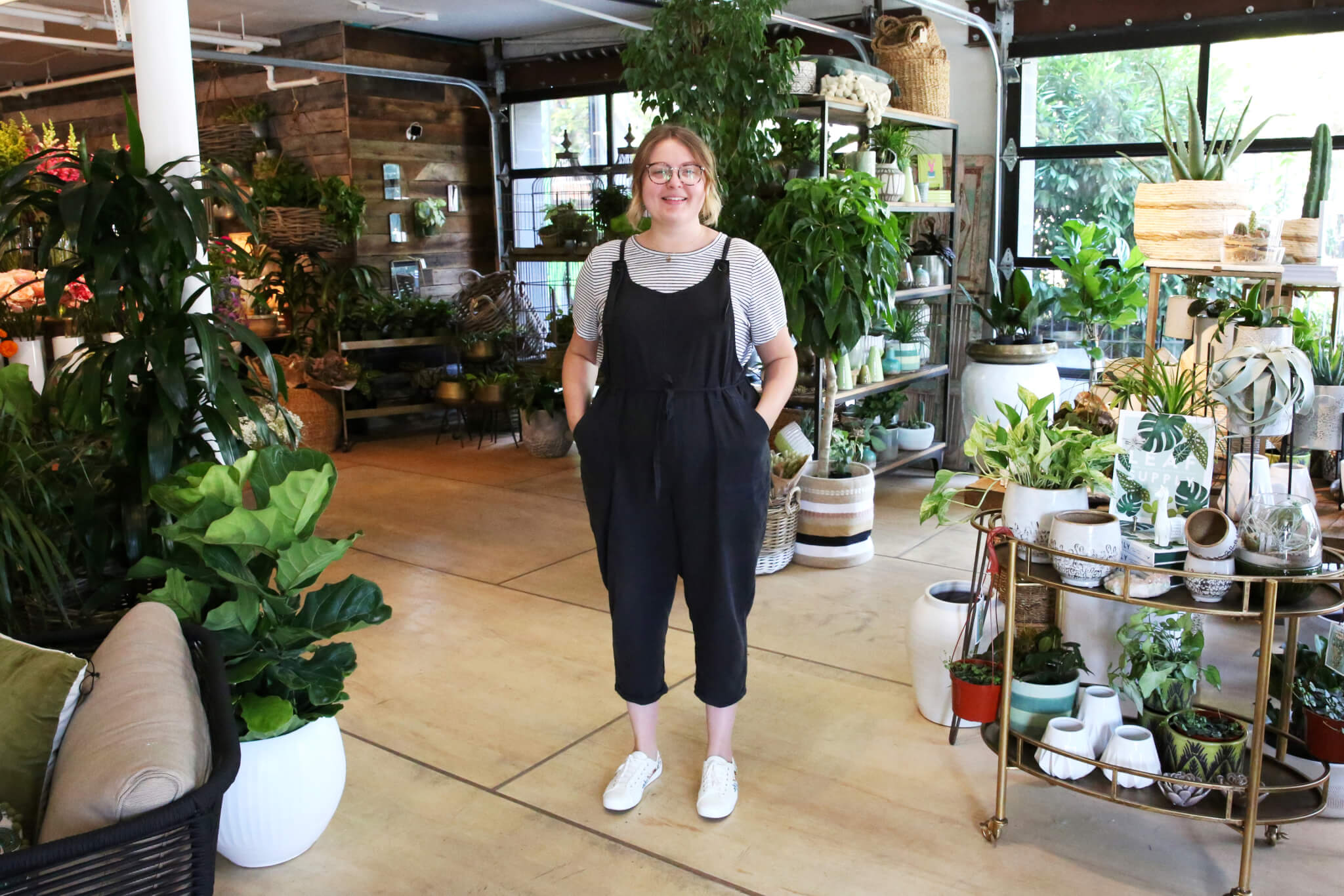 A person wearing a striped shirt and black overalls stands smiling in a bright indoor plant shop filled with a variety of potted plants and shelves displaying greenery and pottery.