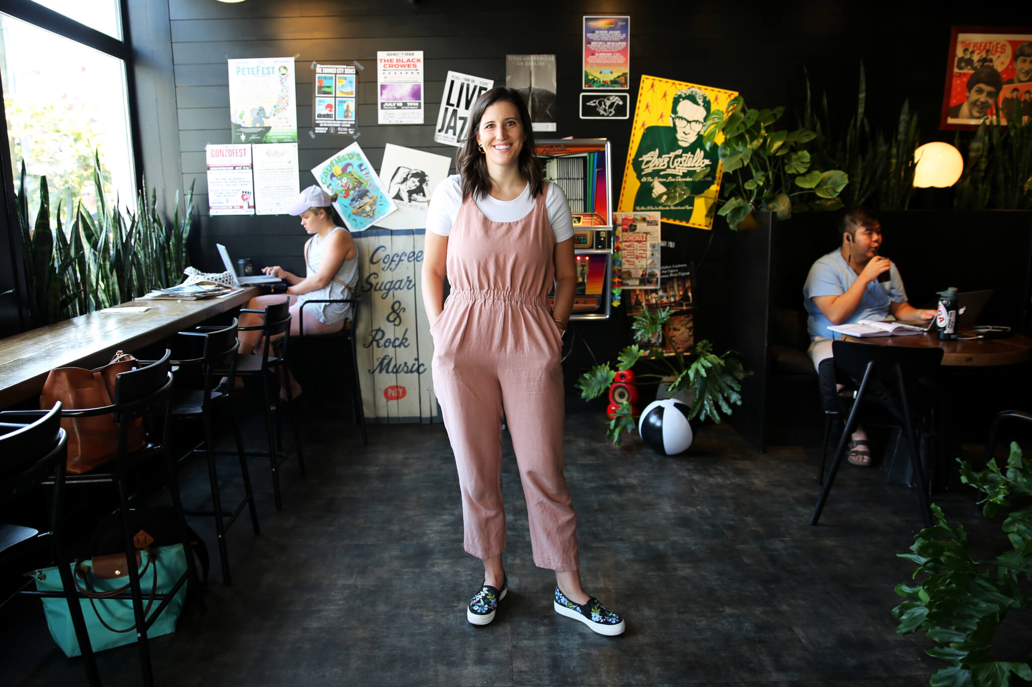 A woman in a pink jumpsuit and white t-shirt stands smiling in a cafe decorated with colorful posters, while other people work on laptops and sit at tables in the background.