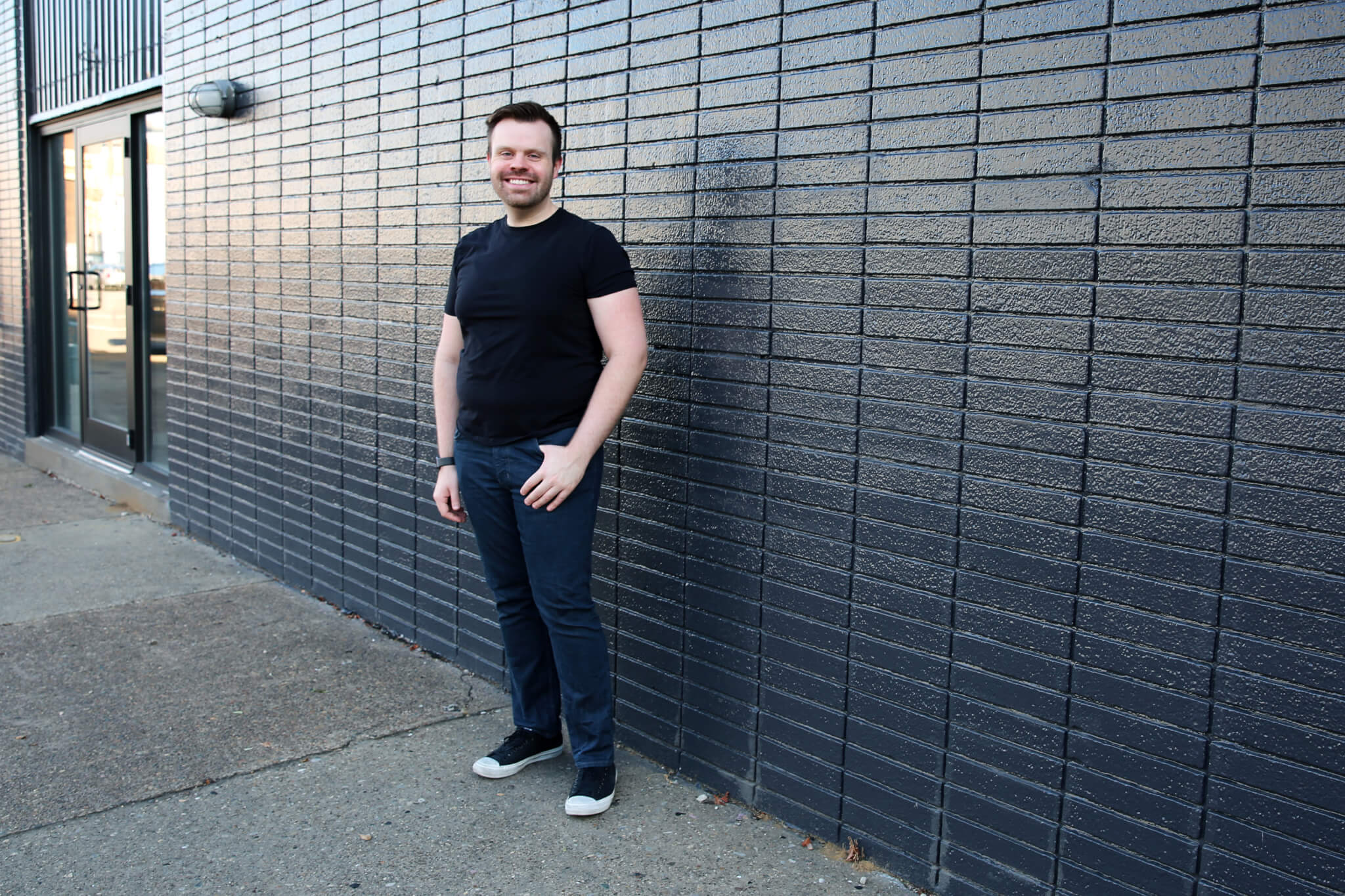 A man in a black t-shirt and jeans stands smiling against a dark brick wall on a sidewalk near a building entrance.