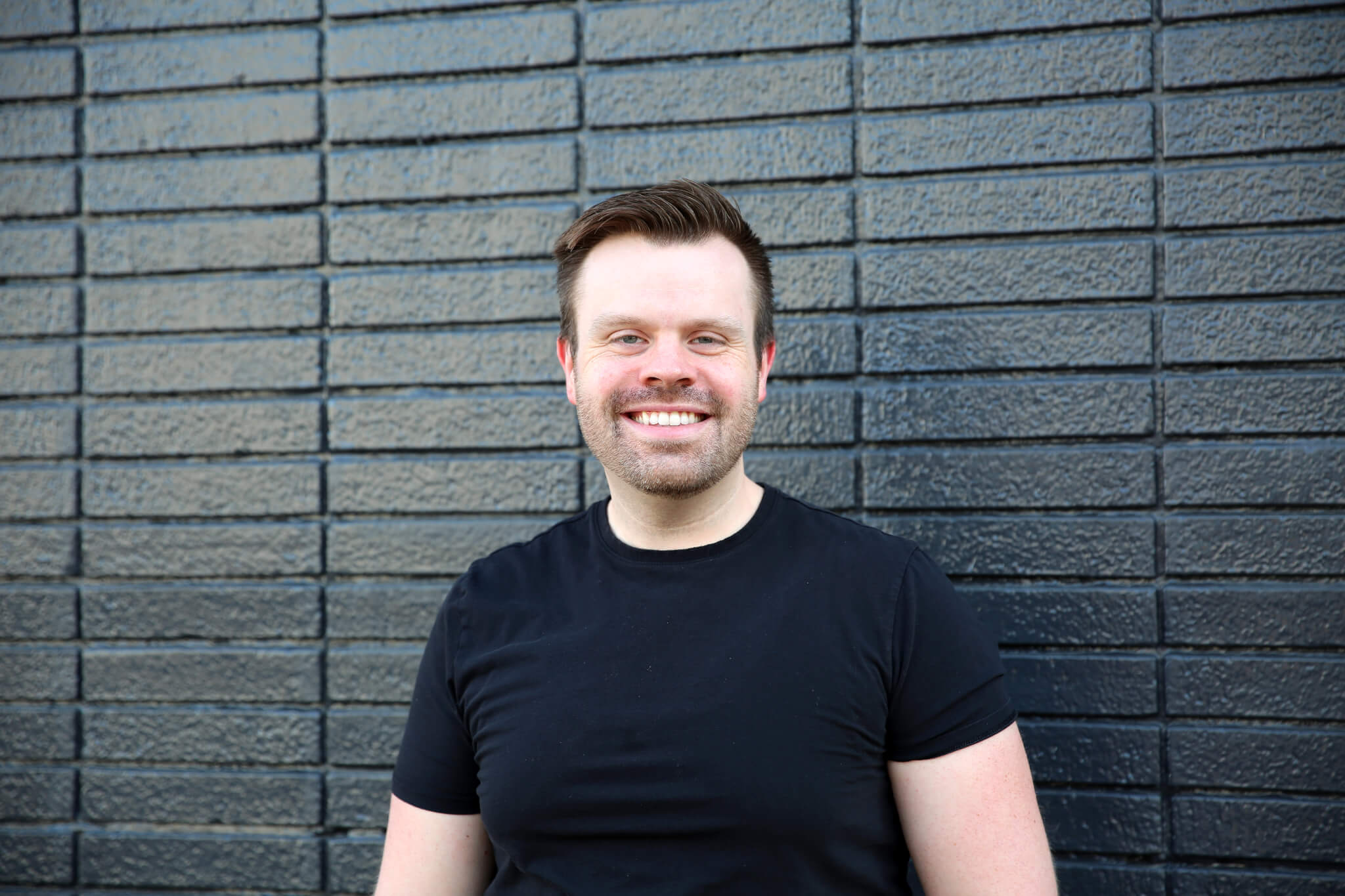 A man with short hair and a beard, wearing a black t-shirt, stands smiling in front of a black brick wall.