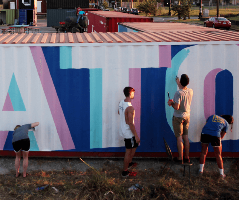 Four people paint large colorful letters on the side of a shipping container during daylight. Three are standing, one is bent over, and various painting supplies are visible on the ground nearby.
