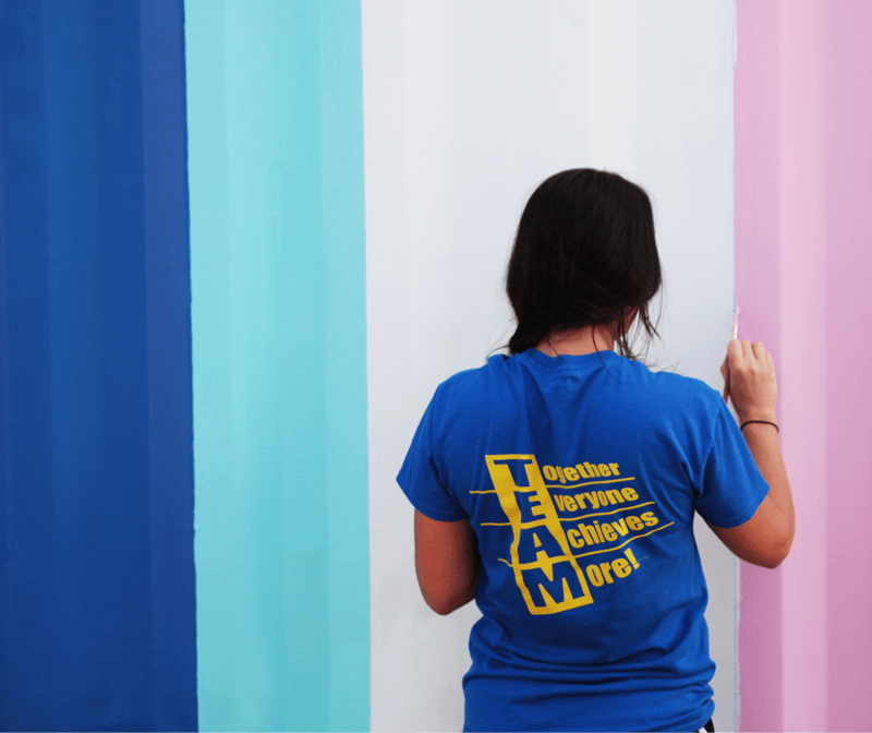 A person wearing a blue TEAM T-shirt is painting vertical stripes in blue, teal, white, and pink on a wall. The text on the shirt reads, Together Everyone Achieves More!.