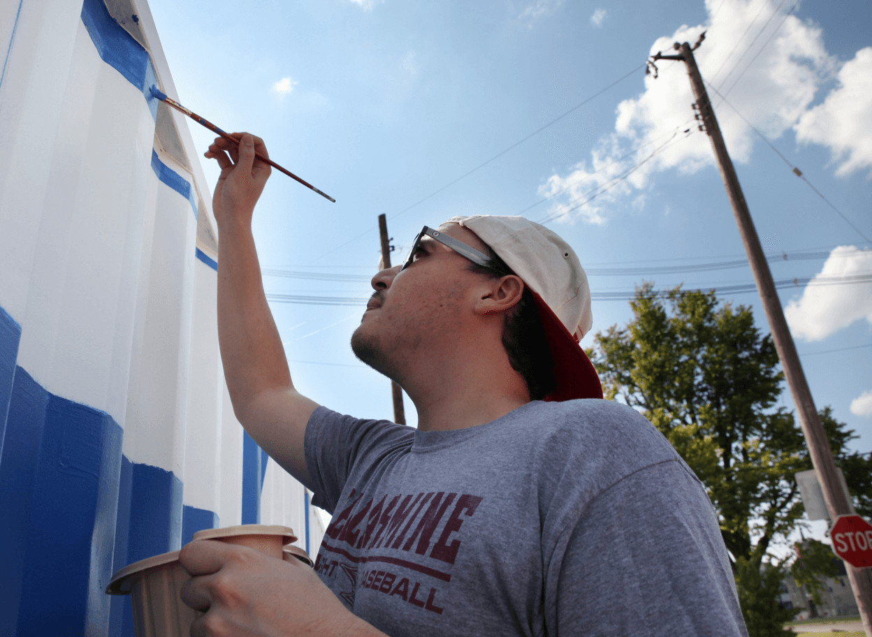 A person wearing a gray T-shirt, sunglasses, and a backward cap paints blue lines on a white wall outdoors, holding a paintbrush in one hand and a cup in the other under a partly cloudy sky.