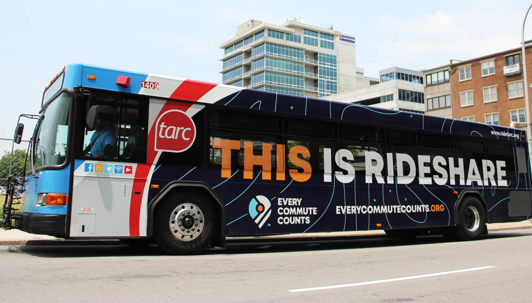 A city bus with a blue and black design is parked on the street. The side displays the text “THIS IS RIDESHARE” along with the Every Commute Counts logo and website, in front of modern office buildings.