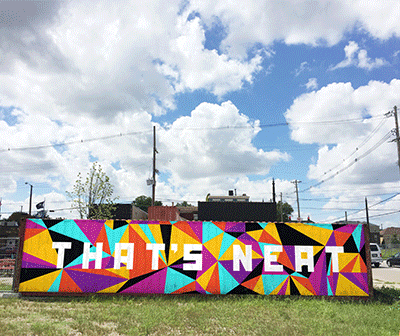 A colorful mural with geometric patterns spells out THATS NEAT in bold white letters against a backdrop of grass, buildings, and a partly cloudy sky.