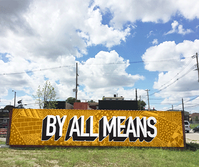 A large outdoor mural reads BY ALL MEANS in bold white letters on a yellow patterned background, set in an open grassy area under a partly cloudy sky.
