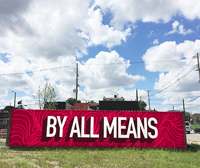 A large outdoor sign with a red patterned background displays the bold white text BY ALL MEANS against a backdrop of grass, power lines, buildings, and a partly cloudy sky.