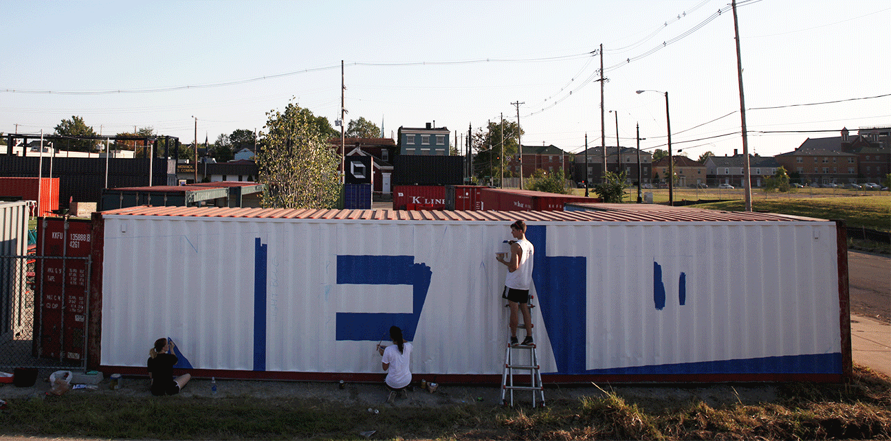 Three people are painting a large, white shipping container with blocks of blue paint in an outdoor, urban area with power lines, buildings, and grassy lots in the background.