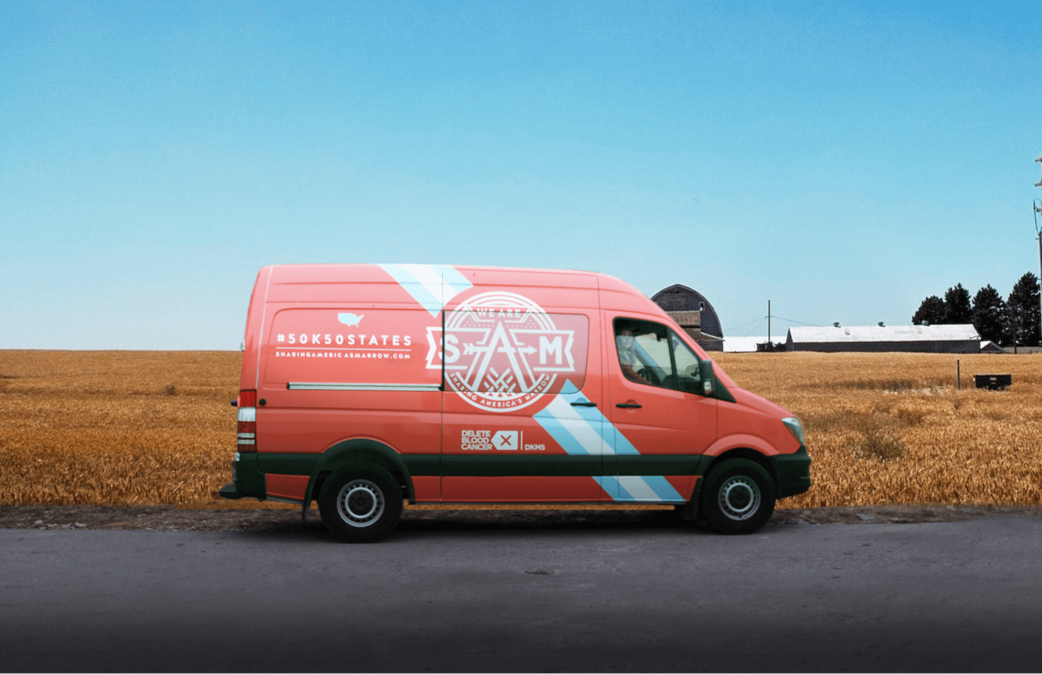A pink and blue van with the Snack Attack logo and #50k50states parked on a rural road beside a golden field, with a barn and trees in the background under a blue sky.