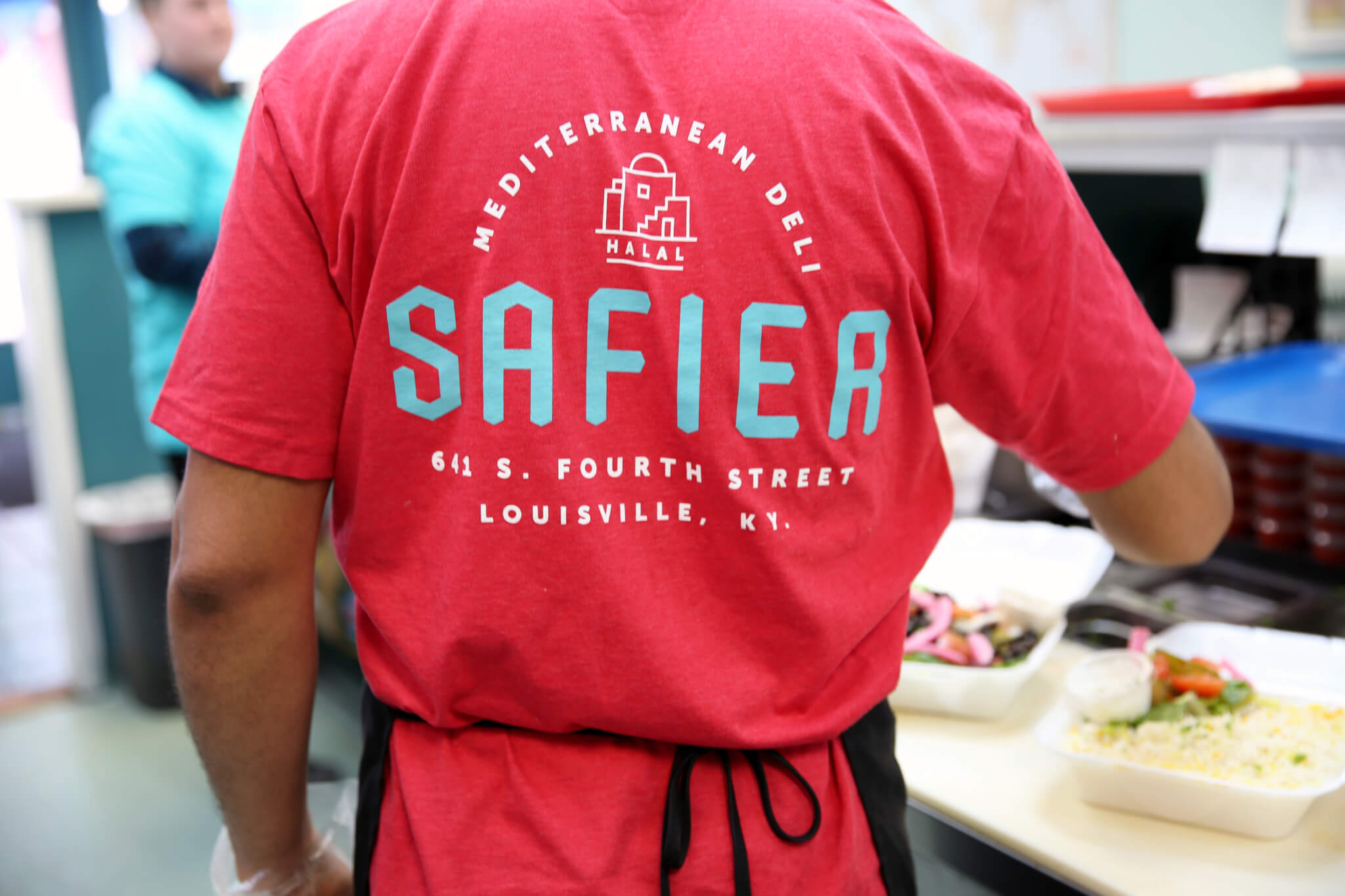 A person wearing a red shirt with Safier Mediterranean Deli Halal, 641 S. Fourth Street, Louisville, KY printed on the back stands near a counter with food containers.