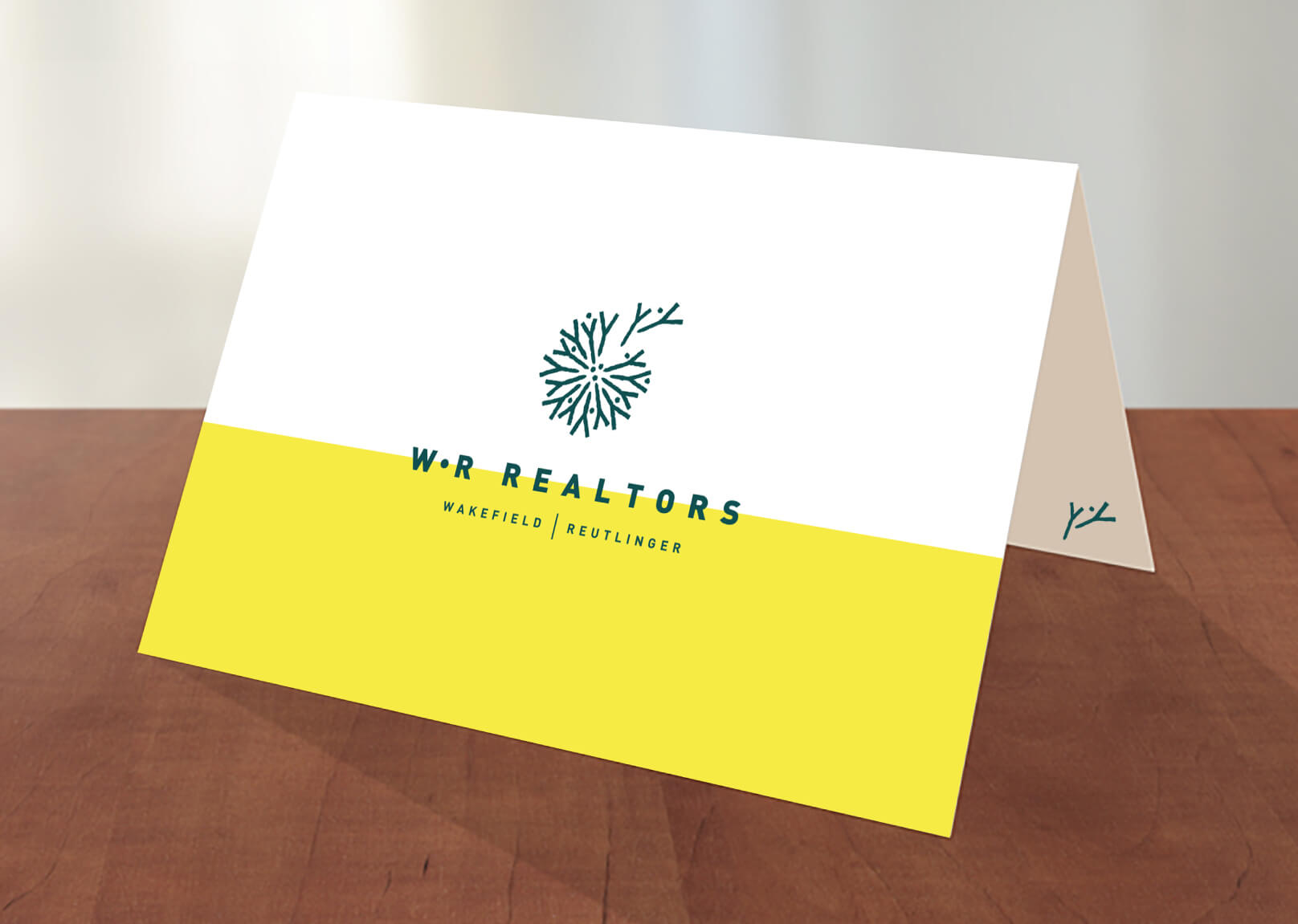 A folded card with a white and yellow design, featuring a green abstract dandelion logo and the text W+R REALTORS WAKEFIELD | REUTLINGER on the front, displayed on a wooden surface.