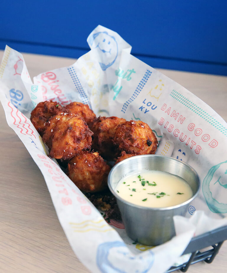 A basket of golden-brown fried balls served on colorful printed paper, accompanied by a small metal cup of creamy dipping sauce garnished with herbs.