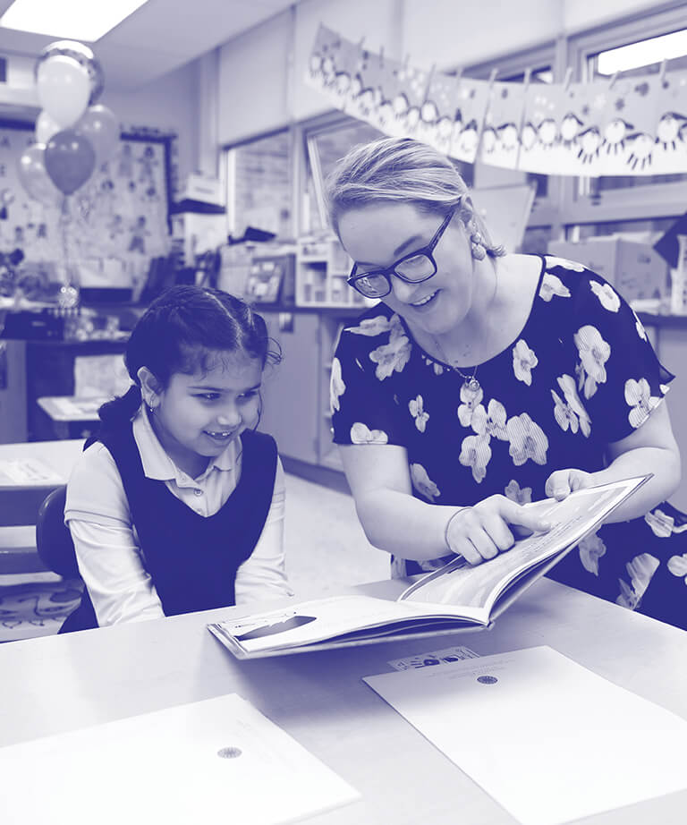 A teacher and a young student sit at a table in a classroom, smiling and looking at an open book together. The teacher is pointing at the book, and classroom decorations are visible in the background.