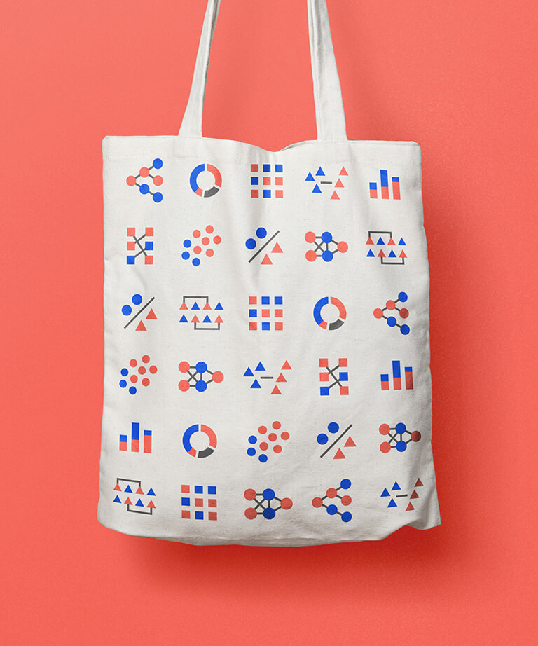 A white tote bag with a pattern of colorful geometric shapes, such as circles, triangles, and squares, in blue, red, and black, arranged in rows against a coral background.