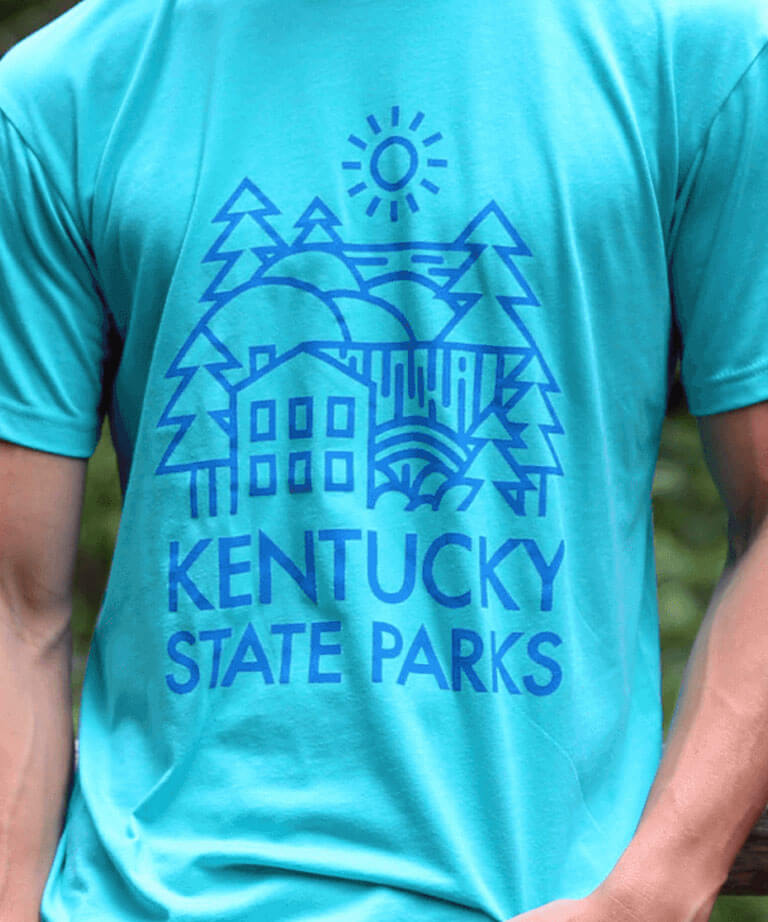 Person wearing a turquoise T-shirt with a blue graphic showing trees, a house, hills, and a sun, along with the text “KENTUCKY STATE PARKS.”.