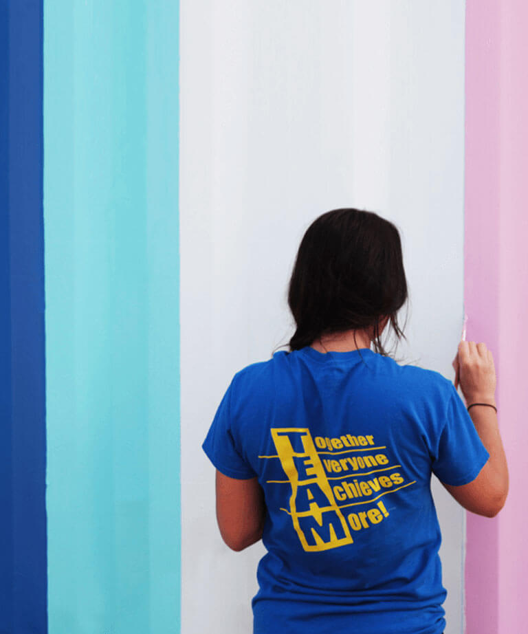 A person with dark hair, wearing a blue shirt that says TEAM: Together Everyone Achieves More!, paints a wall with vertical stripes in blue, white, and pink shades. Their back faces the camera.