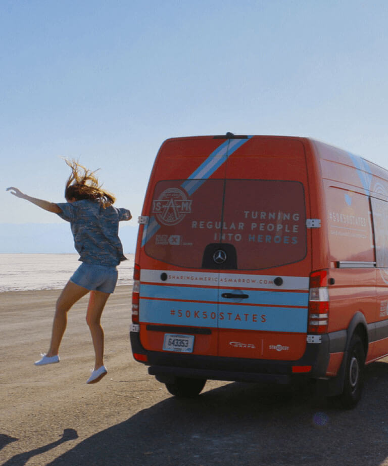 A person in casual clothes jumps with arms raised beside a red van parked on a wide, flat, sunlit landscape; the van reads Turning regular people into heroes.