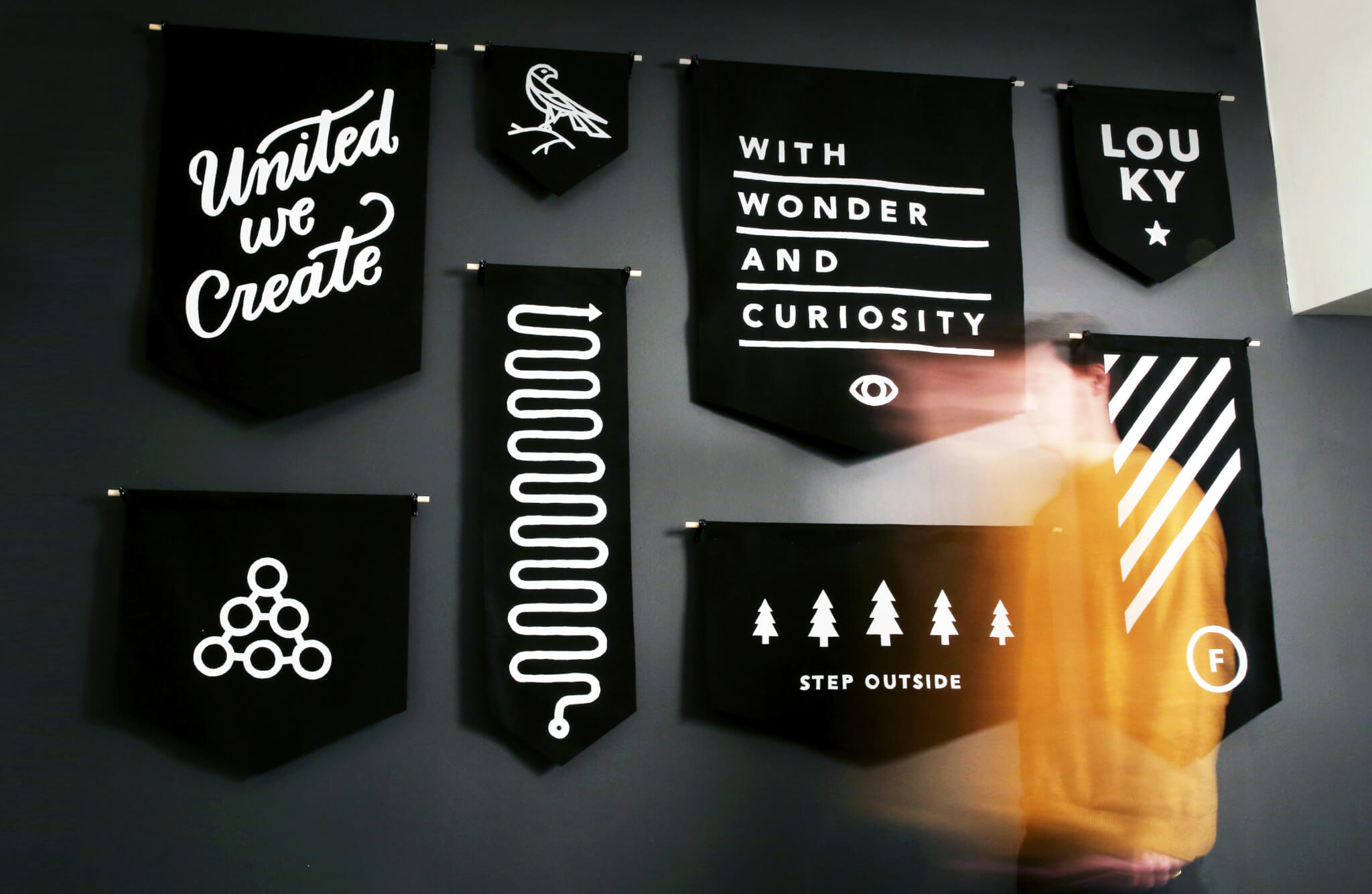 Black banners with white designs and words, including “United we Create,” “With Wonder and Curiosity,” and “Step Outside,” hang on a dark wall. A blurred person in orange passes in the foreground.