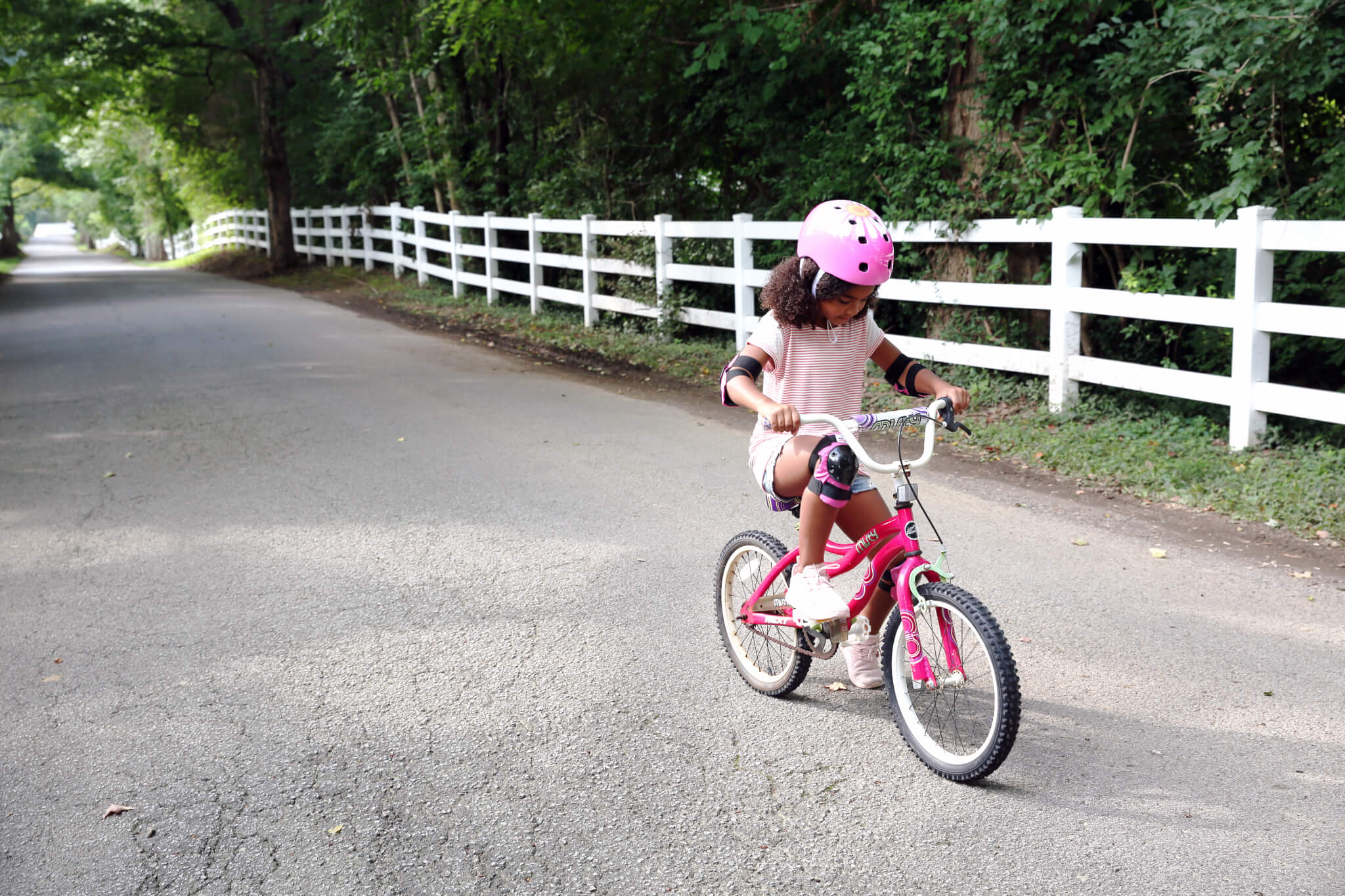 A young girl wearing a pink helmet and protective pads rides a pink bicycle on a paved road lined with a white fence and trees.