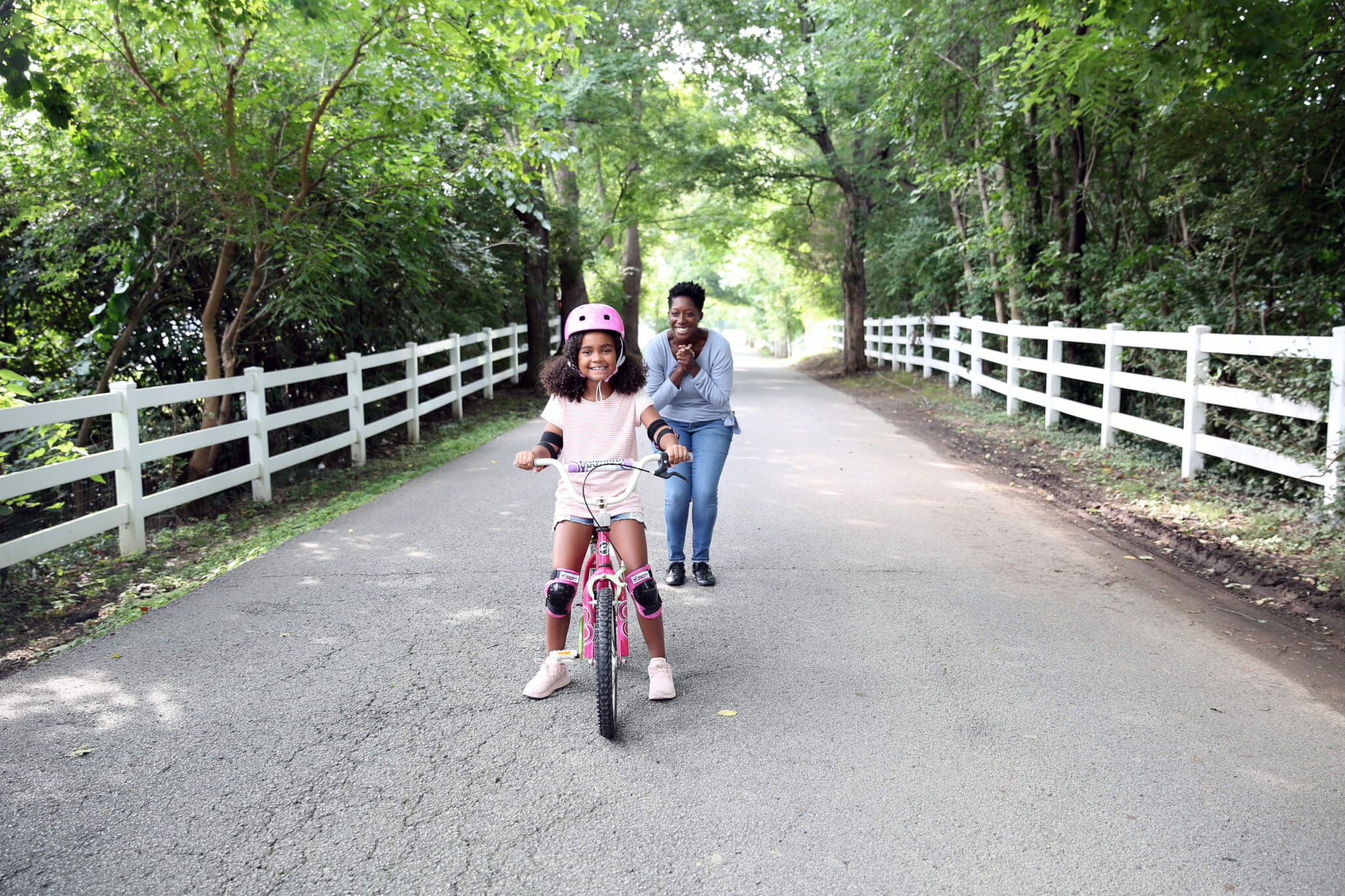 A young girl wearing a helmet and pads rides a pink bicycle on a tree-lined road while an adult walks behind her, smiling and encouraging her. White fences border both sides of the road.
