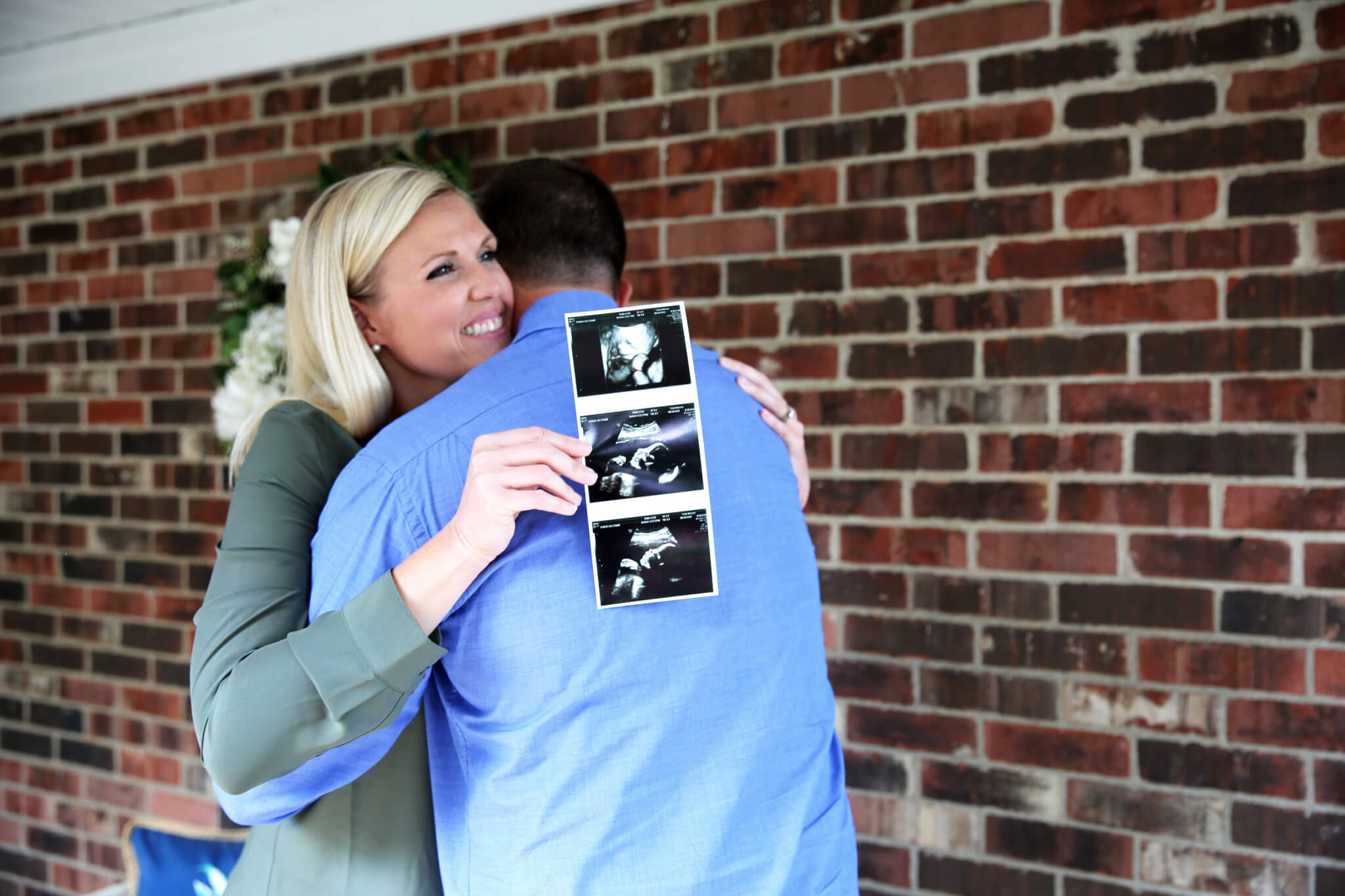 A smiling woman hugs a man while holding up ultrasound photos, standing in front of a brick wall. The image suggests they are happily sharing pregnancy news.