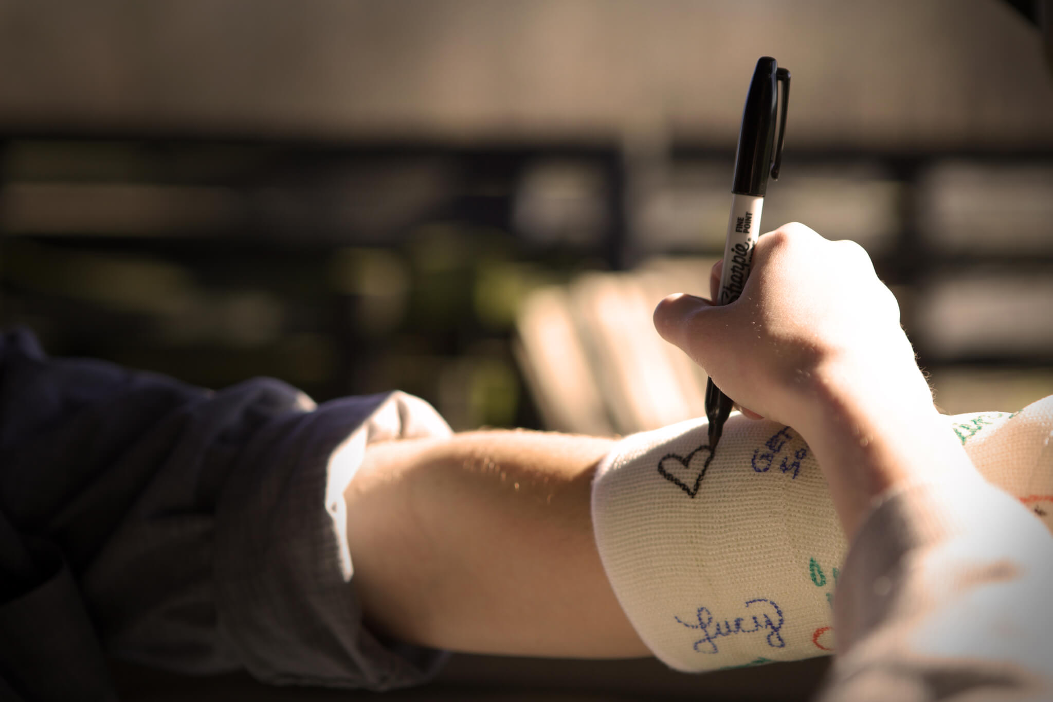A person uses a black marker to draw a heart on a white arm cast that already has several colorful signatures and doodles.