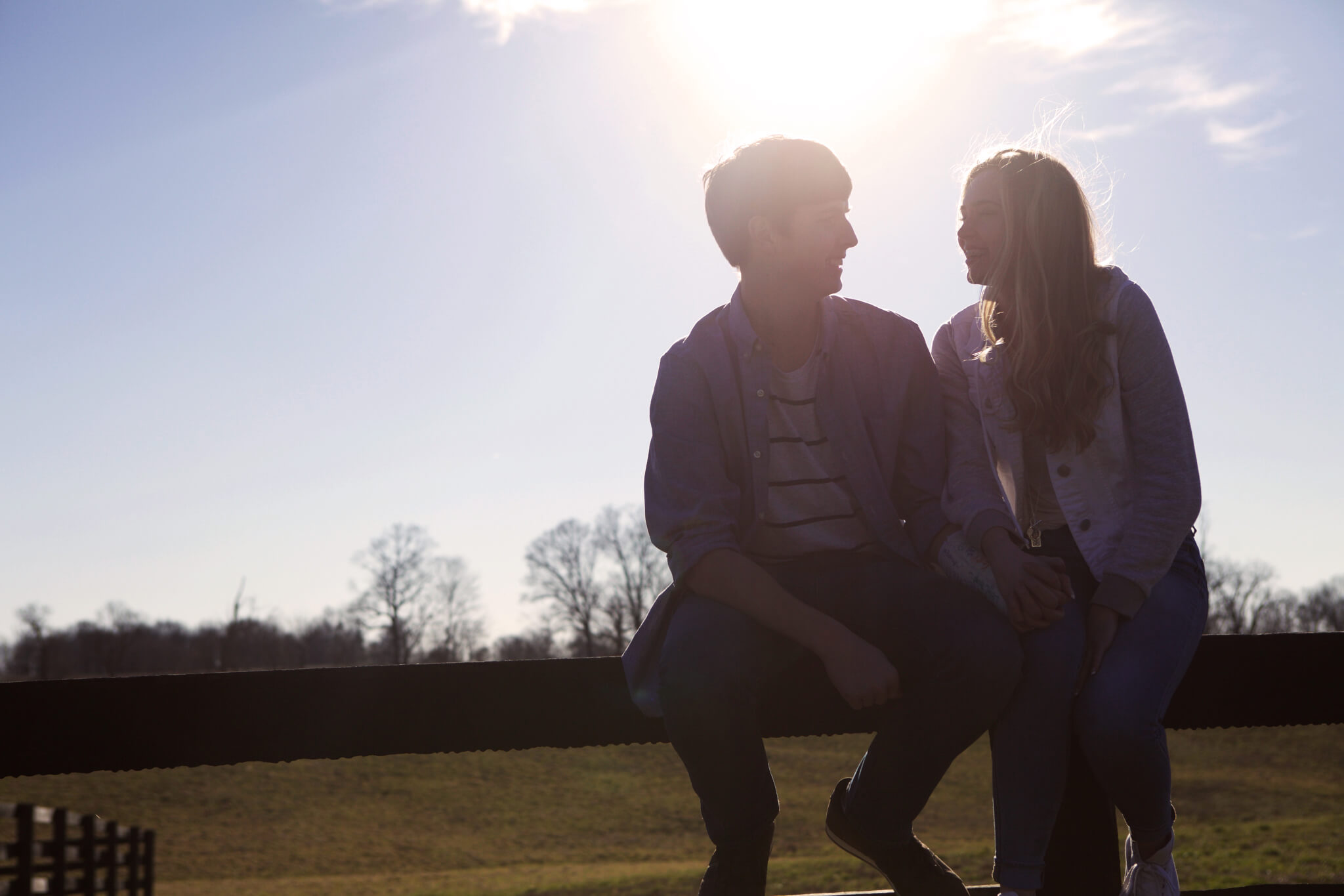 A young couple sits on a fence in a sunlit field, facing each other and smiling, with the bright sun shining overhead and bare trees visible in the background.