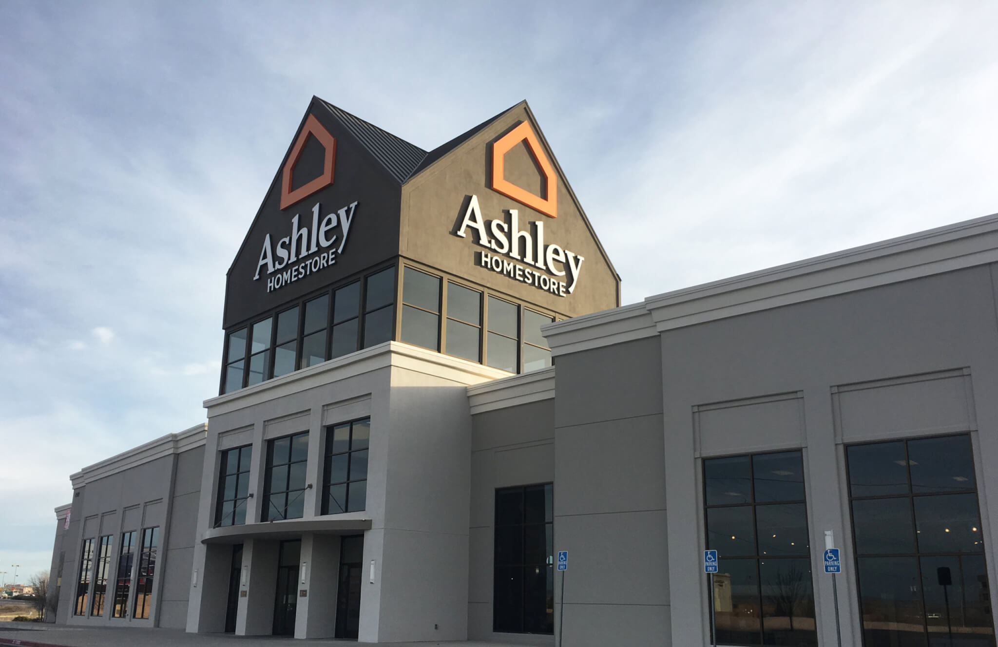 A large Ashley HomeStore building with prominent signage, large windows, and accessible parking spaces in front, set against a partly cloudy sky.