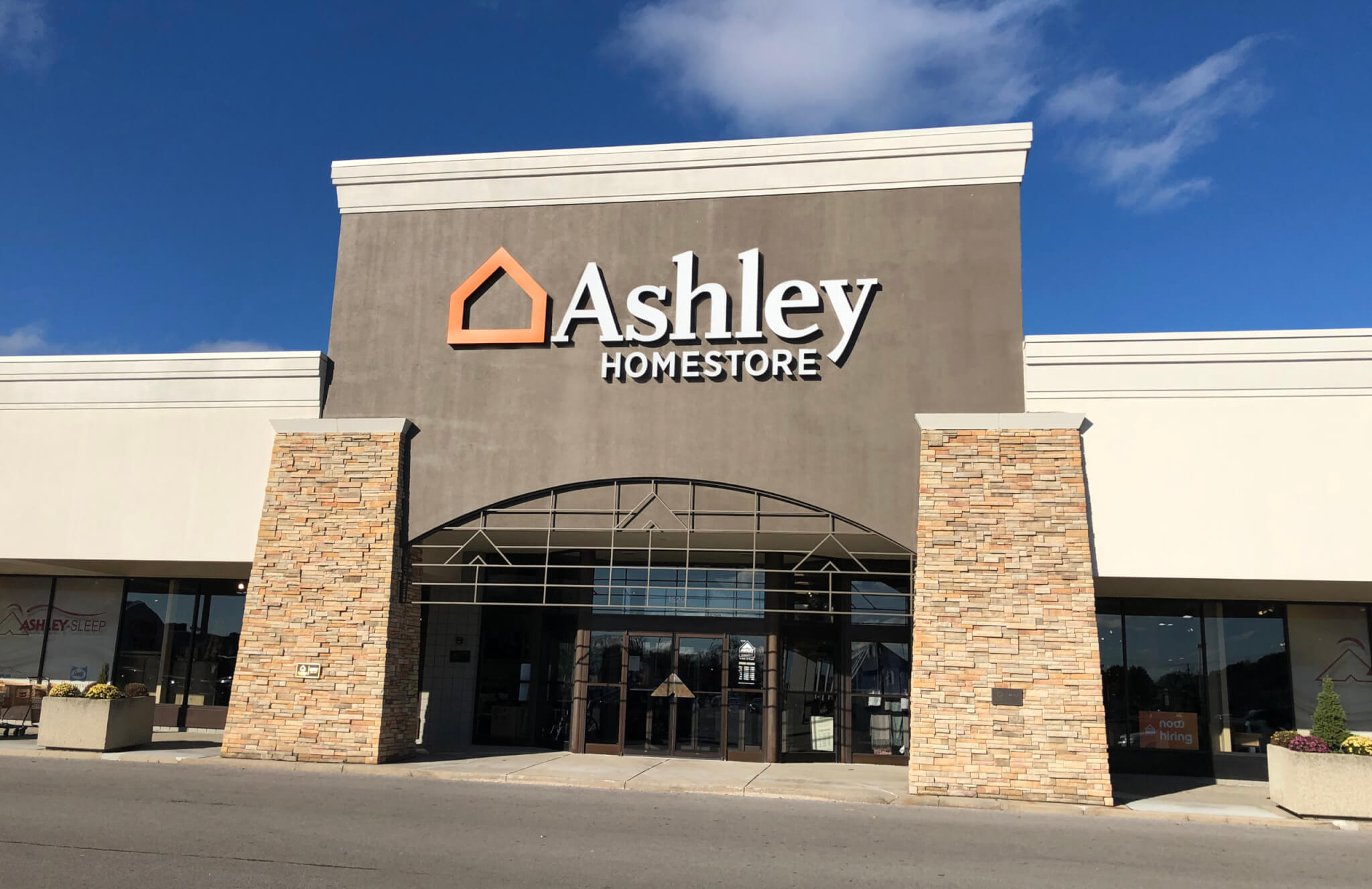 Front view of an Ashley HomeStore retail building with large signage above the entrance, stone pillars, and glass doors under a clear blue sky.
