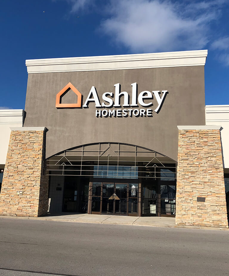 The front entrance of an Ashley HomeStore, featuring large glass doors, stone pillars, and the stores logo above the entrance against a blue sky.