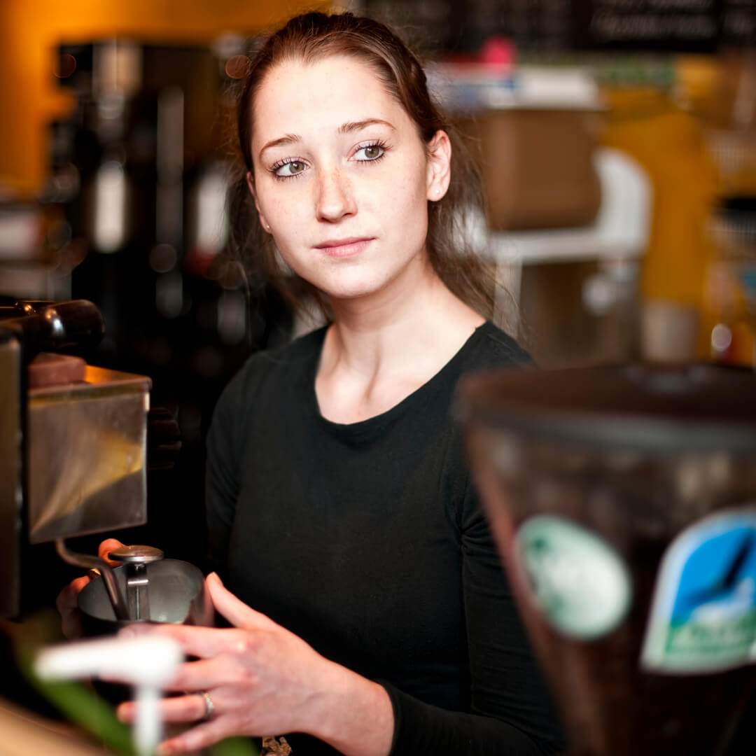 A woman with brown hair tied back, wearing a black shirt, stands behind a coffee machine in a café, holding a portafilter and looking off to the side with a thoughtful expression.
