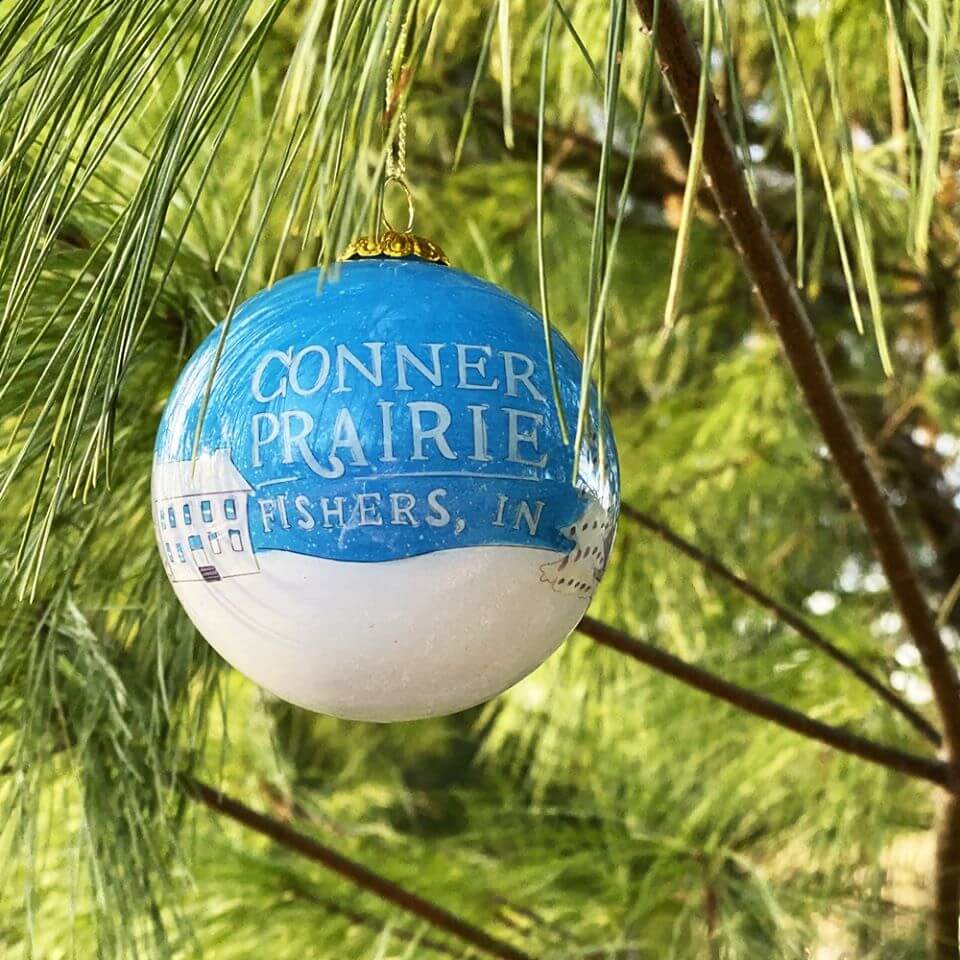 A blue and white Christmas ornament reading Conner Prairie Fishers, IN hangs from a pine tree branch, with green pine needles in the background.