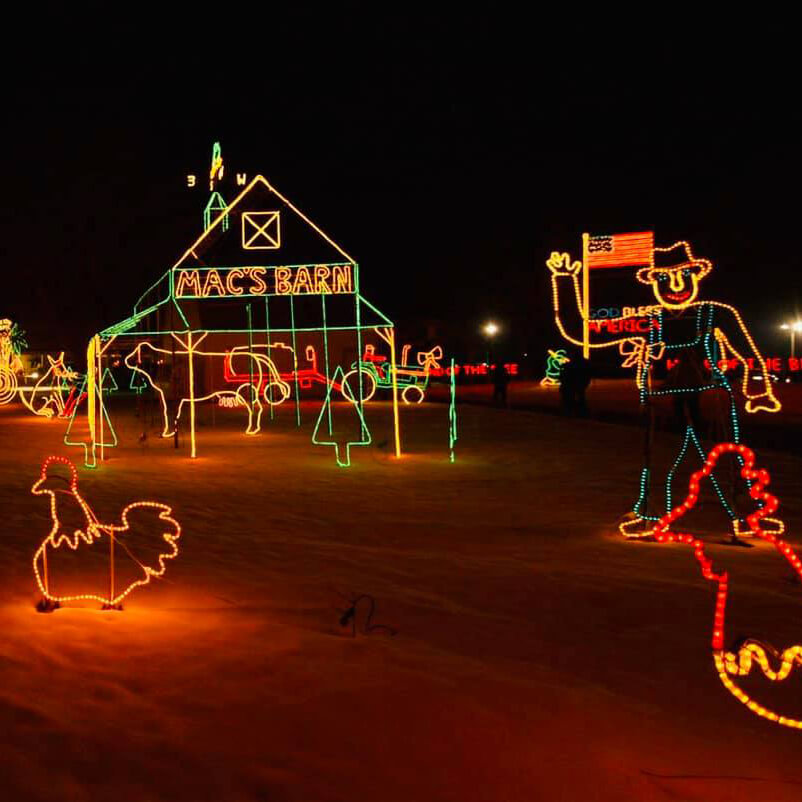 Colorful holiday lights form the shapes of a barn, a rooster, and a cowboy holding an American flag. The barn says MACS BARN, all set against a dark night background.