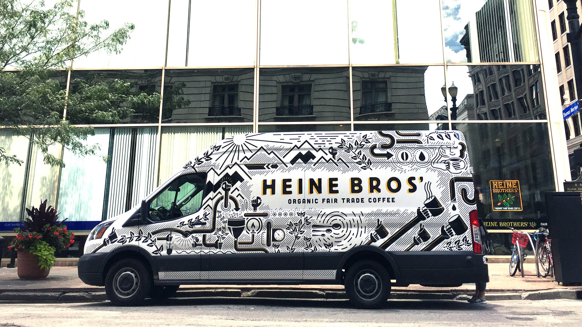 A Heine Bros van decorated with black and white illustrations and the text Heine Bros Organic Fair Trade Coffee is parked on a city street in front of a glass building.