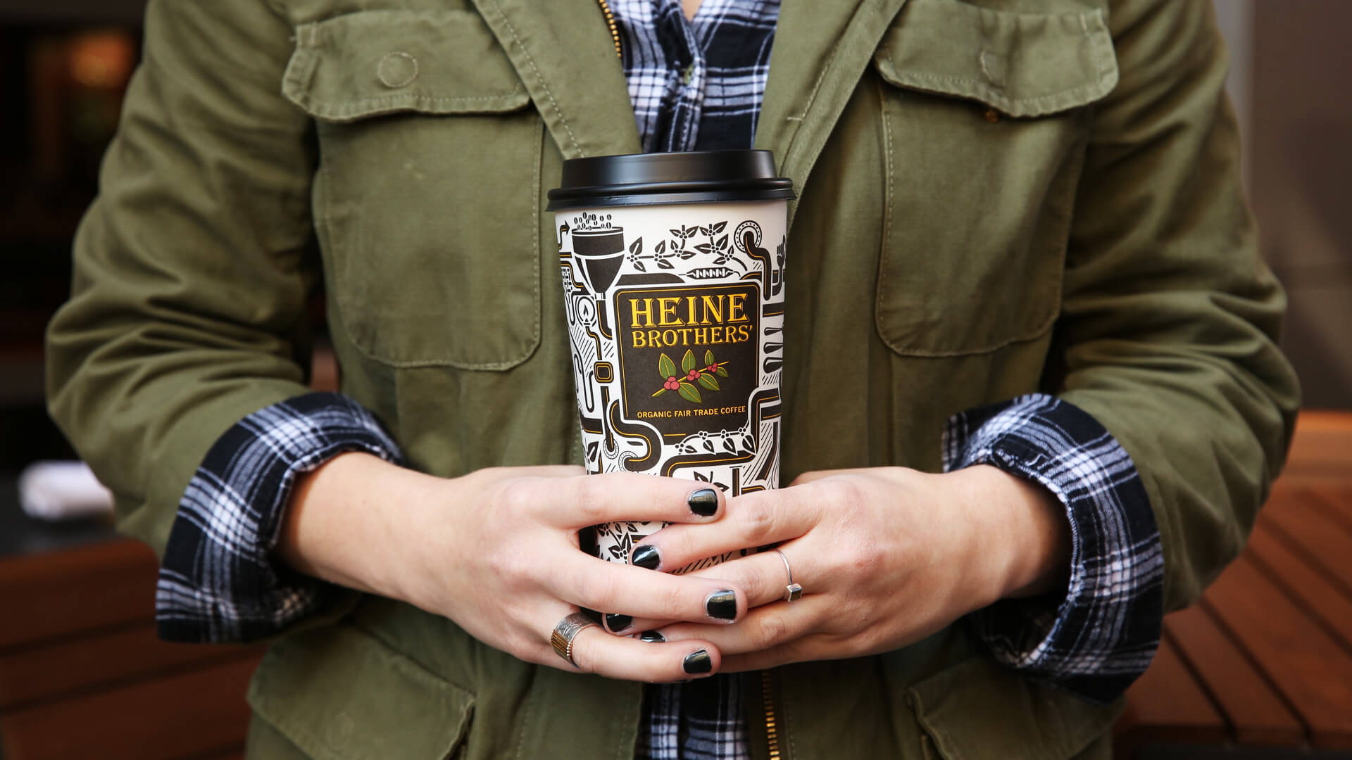 A person wearing a green jacket over a plaid shirt holds a Heine Brothers coffee cup with both hands. The cup has a decorative black-and-white pattern and a black lid. The persons nails are painted black.