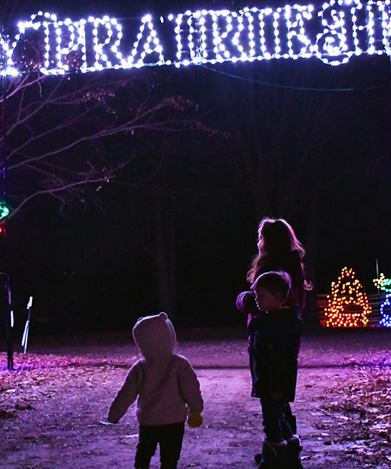 Three children walk under illuminated holiday lights spelling PRAIRIE at night, surrounded by trees and colorful light displays in the background.