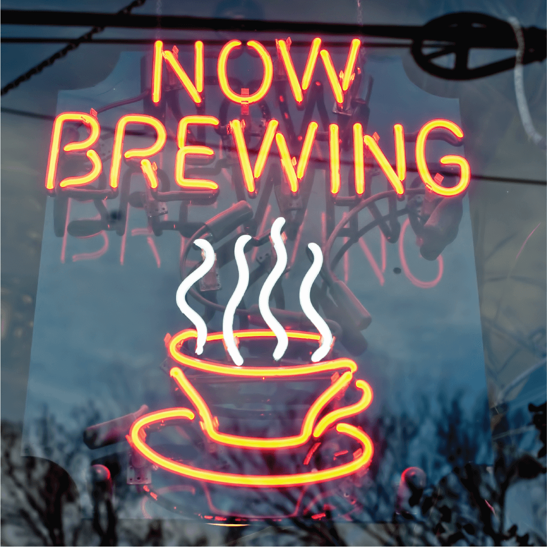 A neon sign with the words NOW BREWING glows above a steaming coffee cup design. The sign is displayed in a window with reflections of trees and power lines visible.