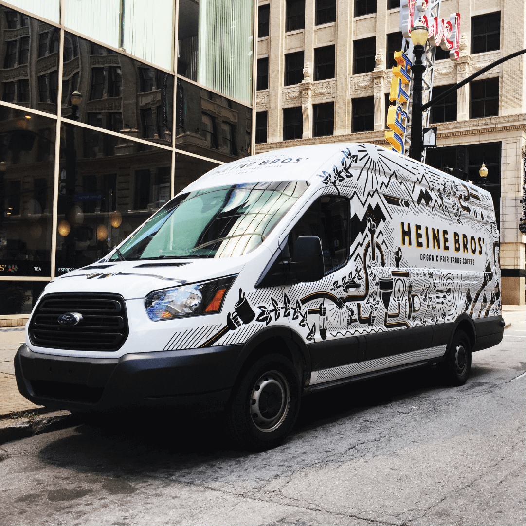 A white Ford van with “Heine Bros. Organic Fair Trade Coffee” and black artistic graphics on its side is parked on a city street in front of tall buildings.
