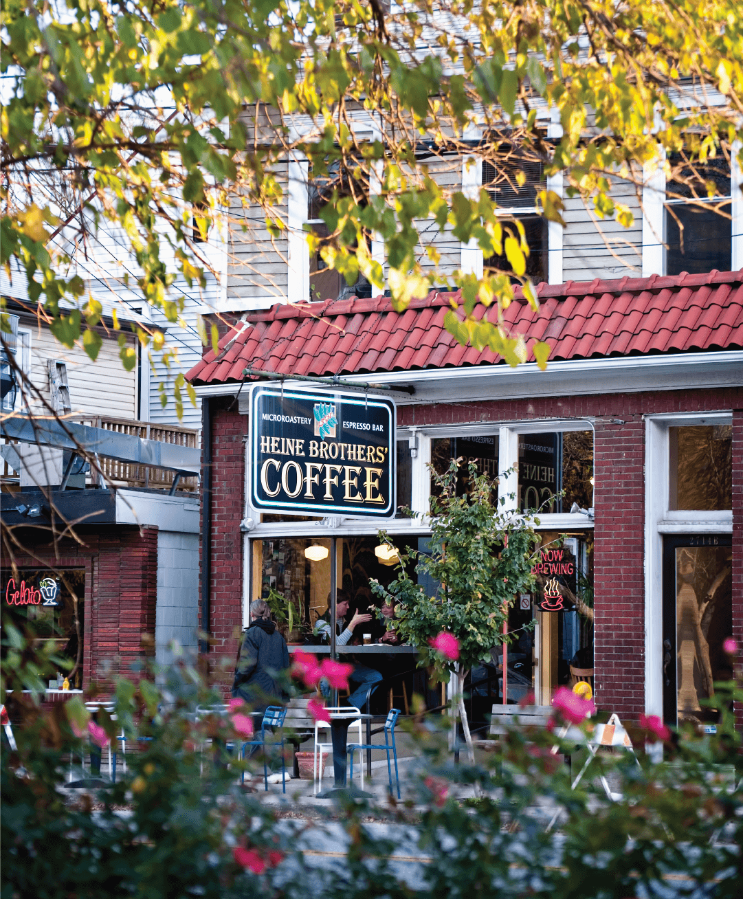 A brick building with a red tile roof houses Heine Brothers Coffee, visible through large windows, surrounded by greenery and outdoor seating on a sunny day.