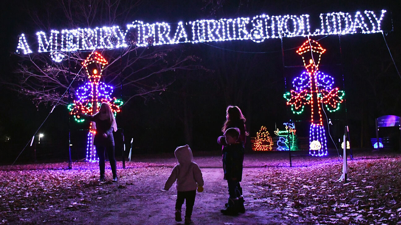 A group of people, including children, walk under a large illuminated sign reading A MERRY PRAIRIE HOLIDAY with colorful holiday light decorations in the background at night.